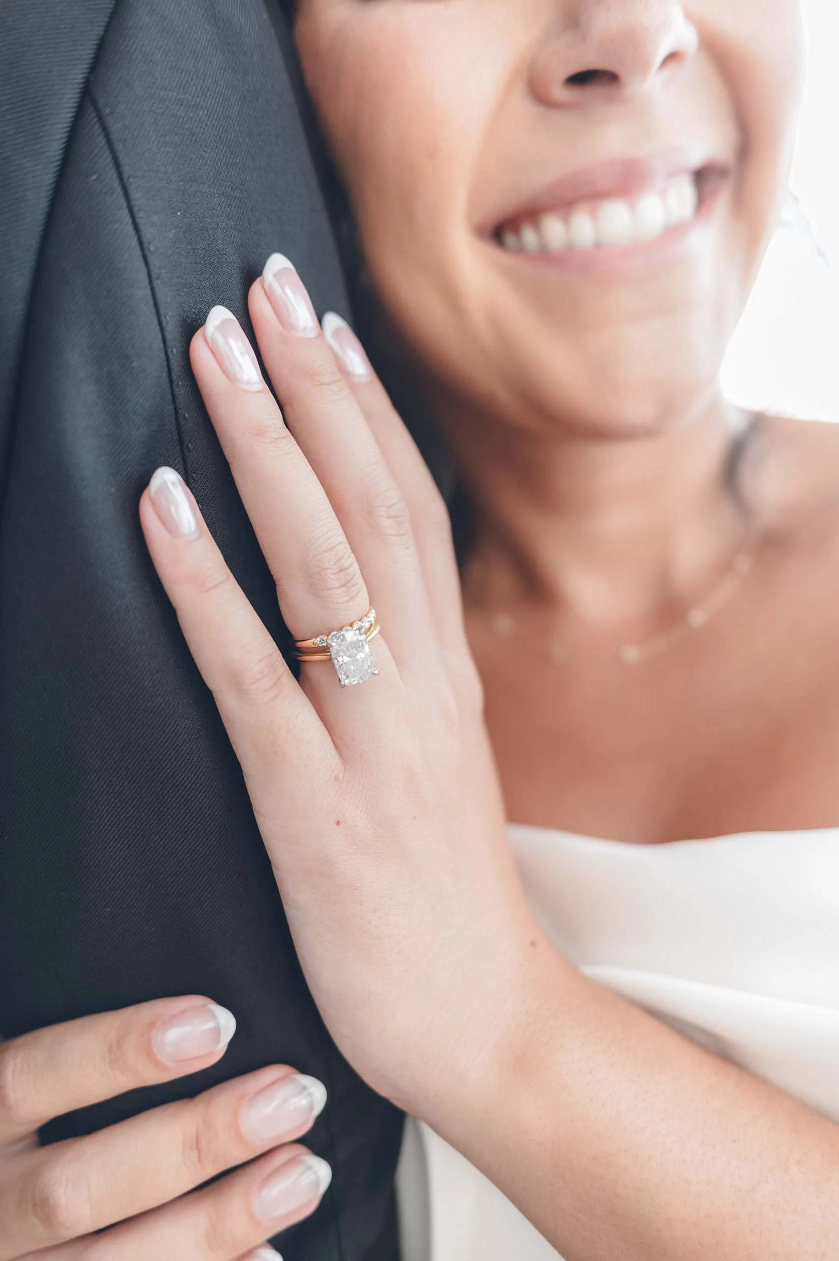 Close-up of a woman with a radiant smile, wearing a diamond engagement ring, and gently holding the arm of the groom.