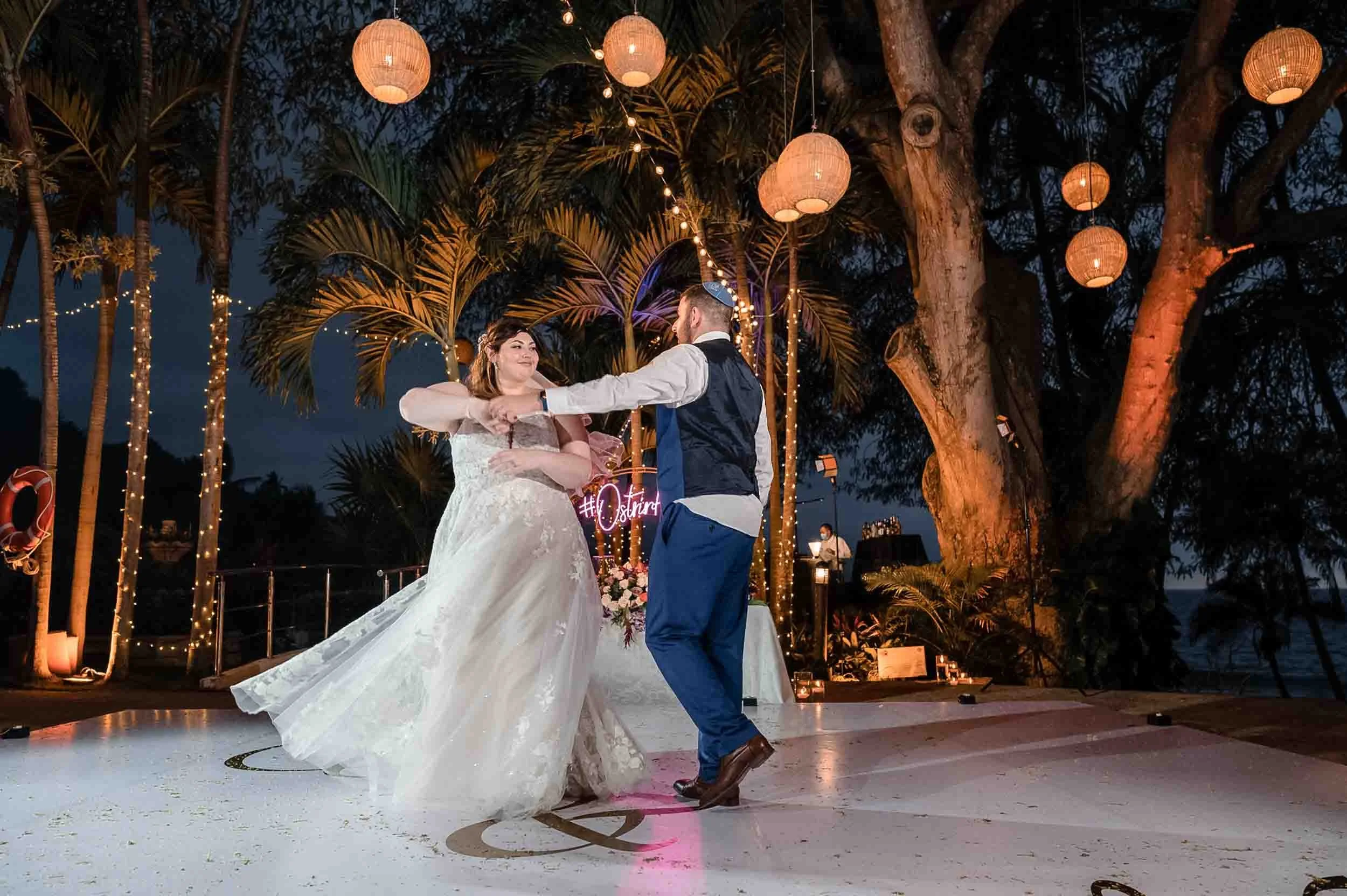 A bride and groom are dancing outside at night during a wedding celebration. The area is decorated with hanging paper lanterns and string lights, set among trees with tropical foliage.
