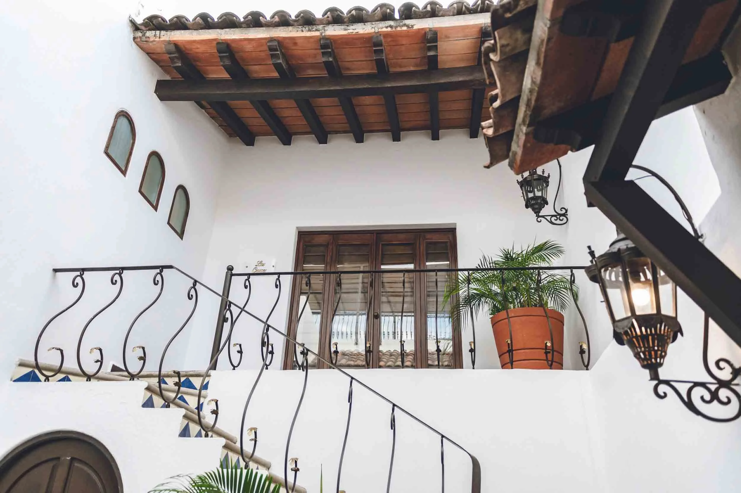 Interior view of a Spanish-style home with a wooden ceiling, white walls, decorative iron railings, and a potted plant near a wooden window.