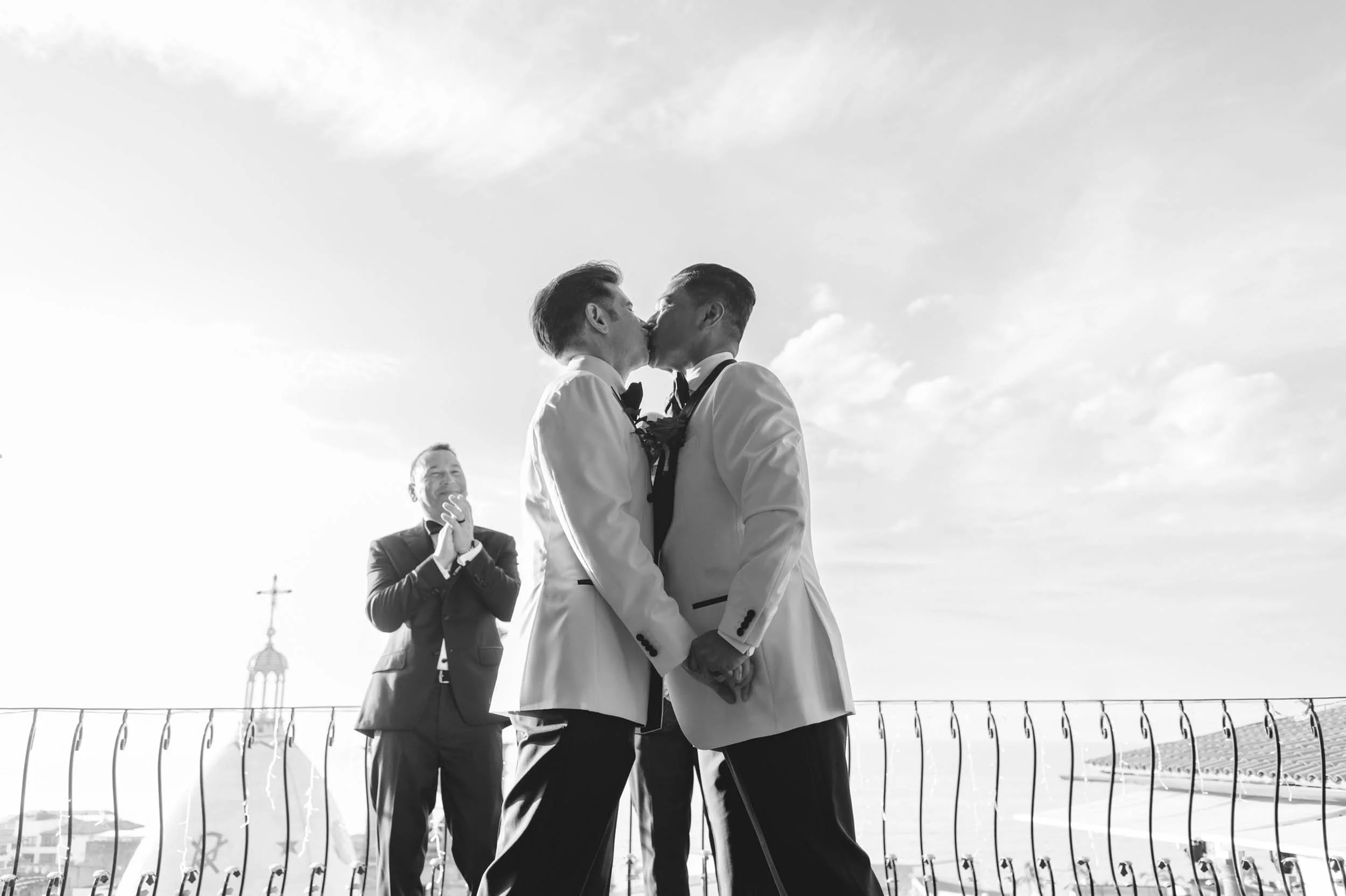 First kiss of gay couple during their wedding ceremony, while the officiant in the background claps in joy.
