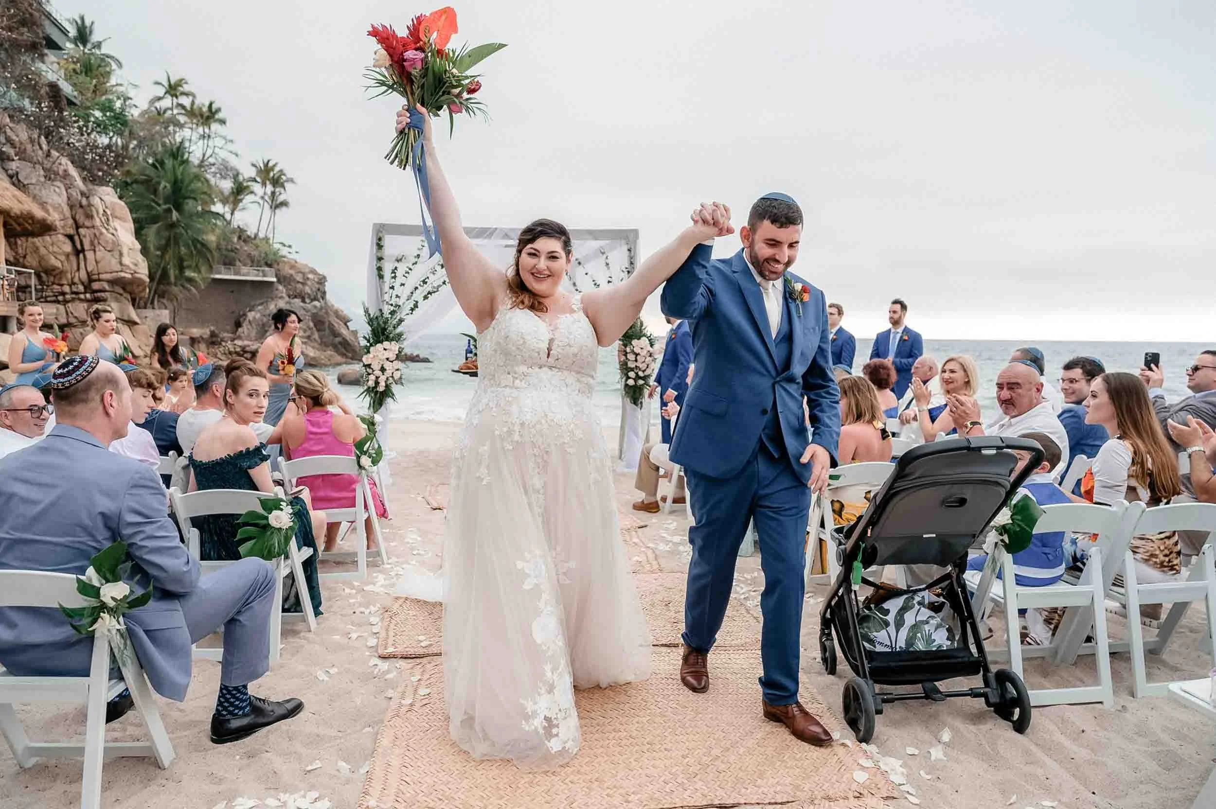 A newlywed couple walking down the aisle at a beach wedding, holding hands and smiling. The bride is wearing a white lace gown and holding a bouquet, while the groom is dressed in a blue suit and kippah. Guests are seated on white chairs decorated wi