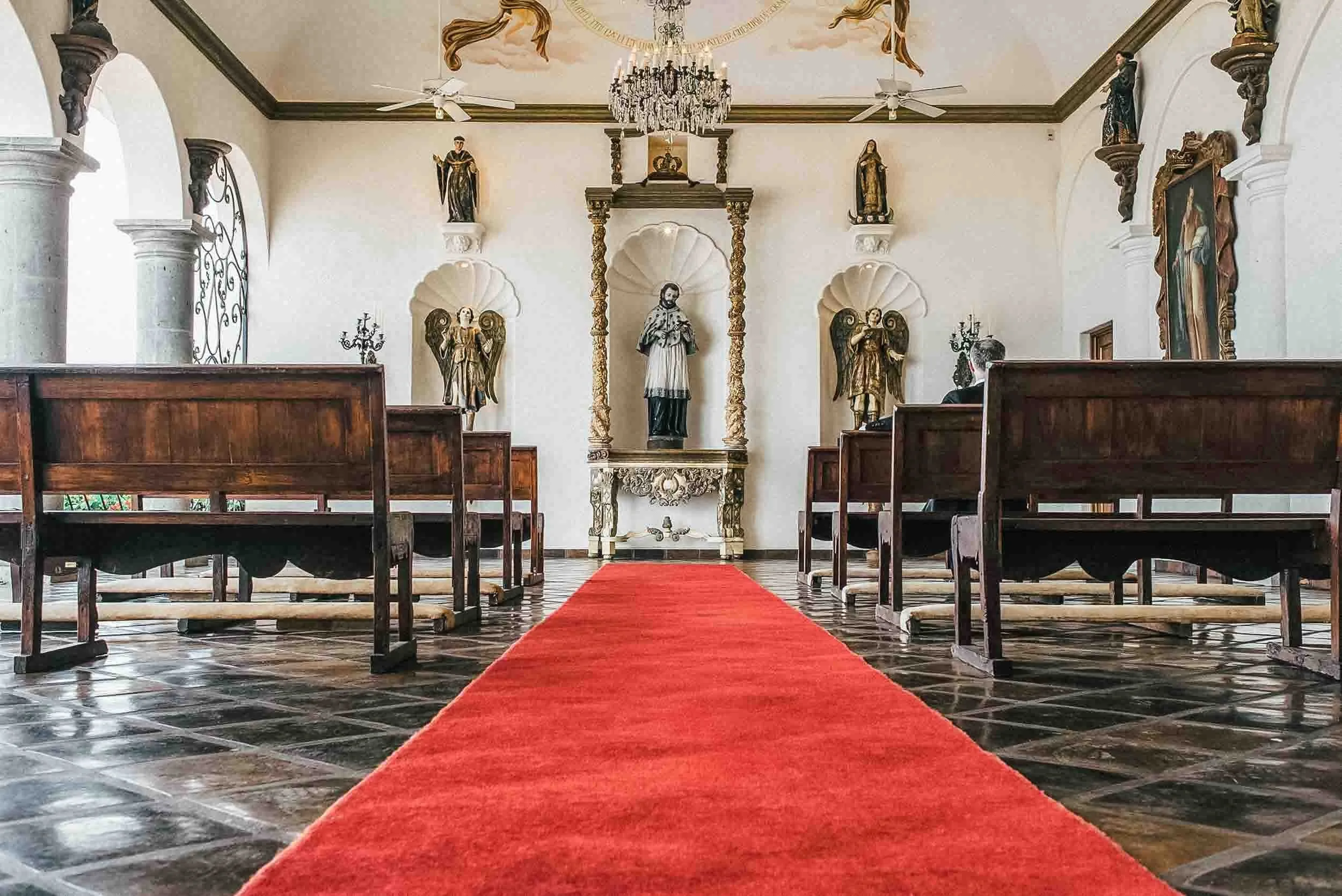 Interior view of La Capella setting with wooden pews, a red carpet leading to an altar with religious statues, chandeliers, and a painted ceiling.