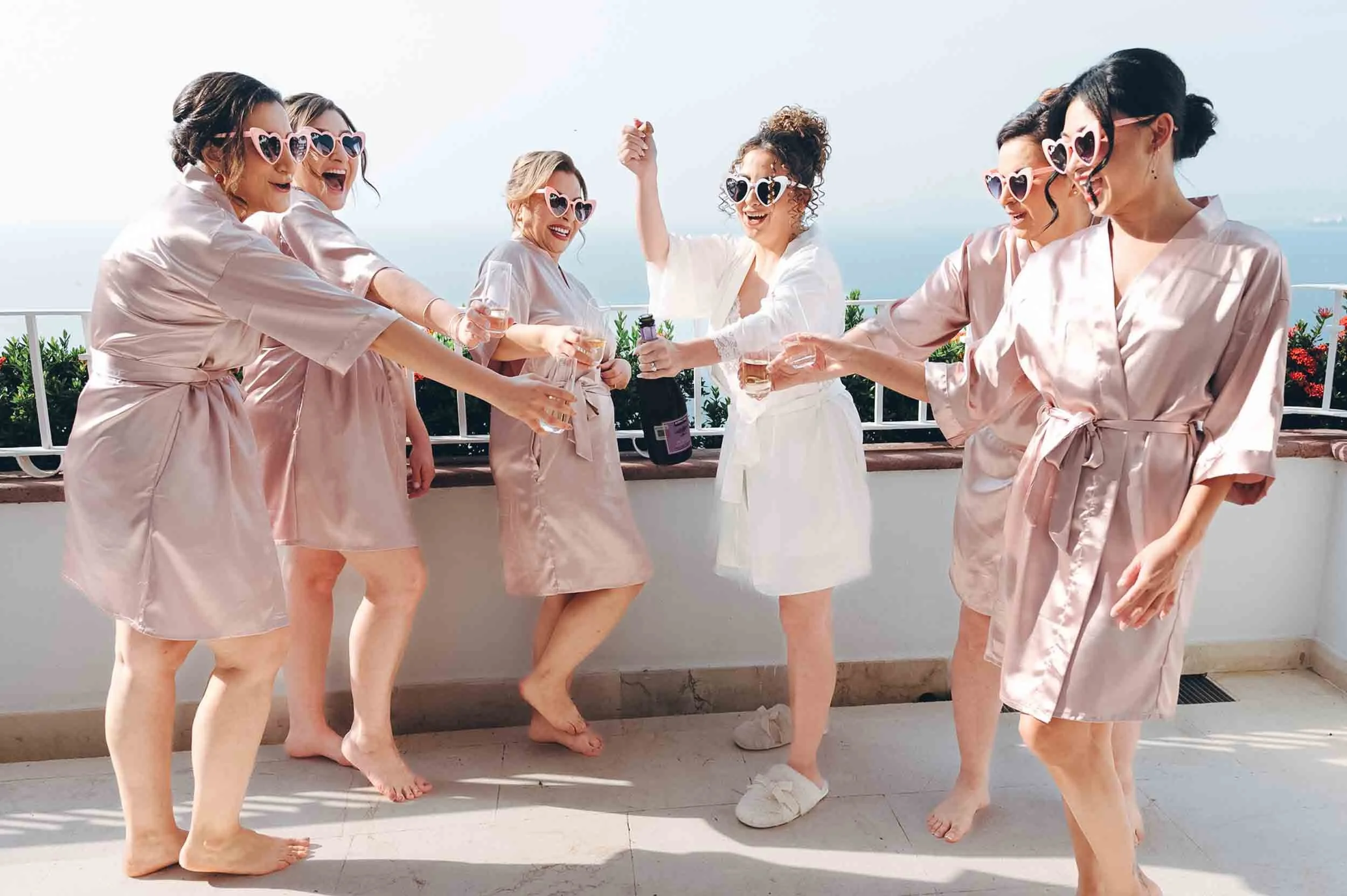 Bride and bridesmaids in satin robes celebrating with drinks on an outdoor terrace, wearing heart-shaped sunglasses, and holding a champagne bottle.
