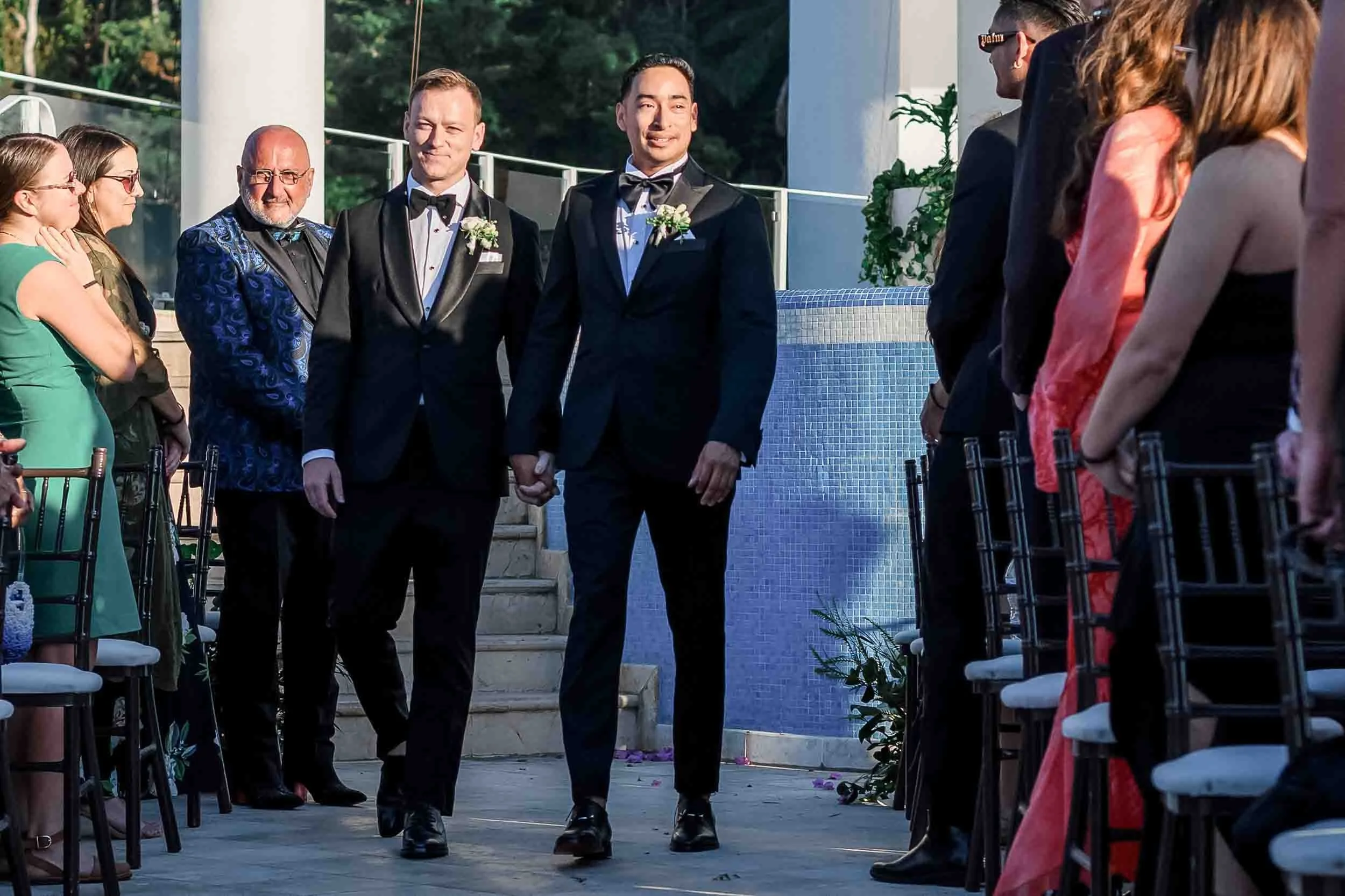 Grooms walking down an aisle at their wedding ceremony, with guests seated on either side, outdoors with a pool and trees in the background.