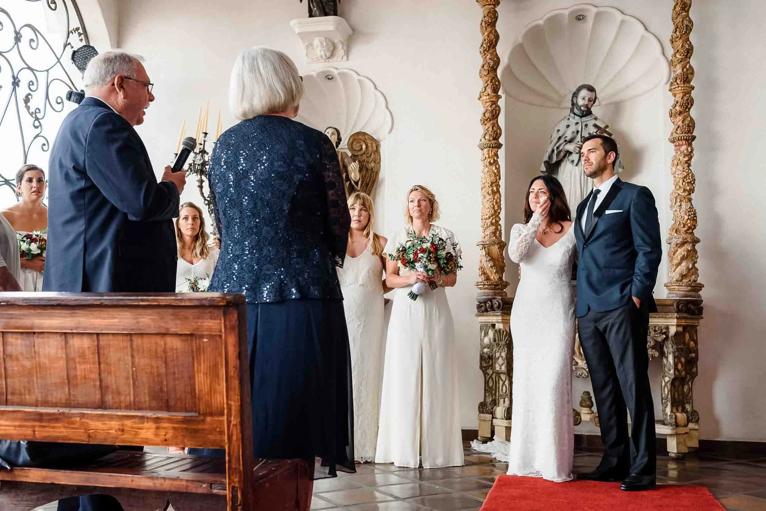 A wedding ceremony with the bride and groom hearing the best wishes of her father, surrounded by women bridesmaids, in a decorated church with statues and ornate columns.