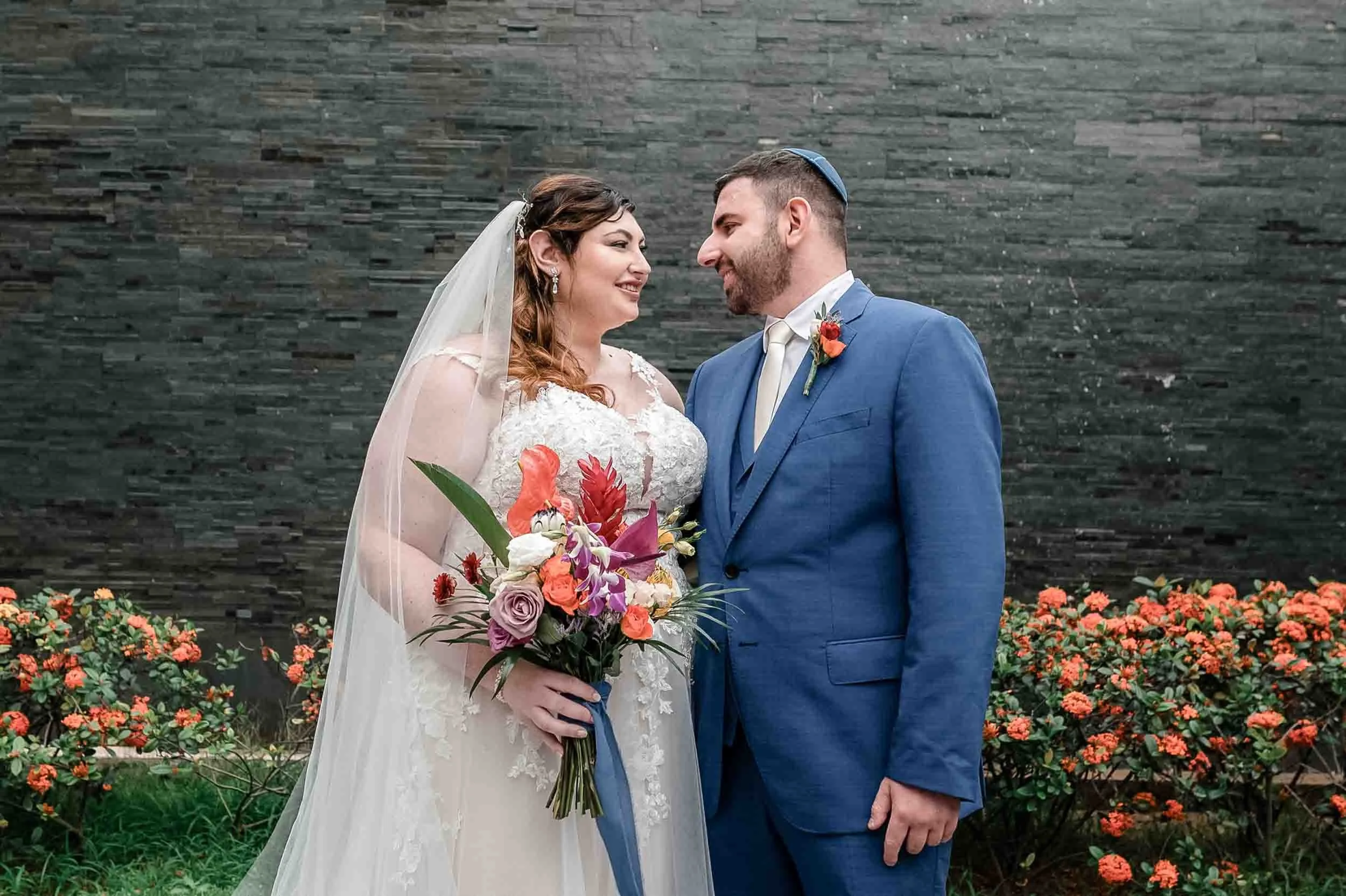A bride and groom portrait stand facing each other, smiling, surrounded by orange and purple flowers, against a dark brick wall.