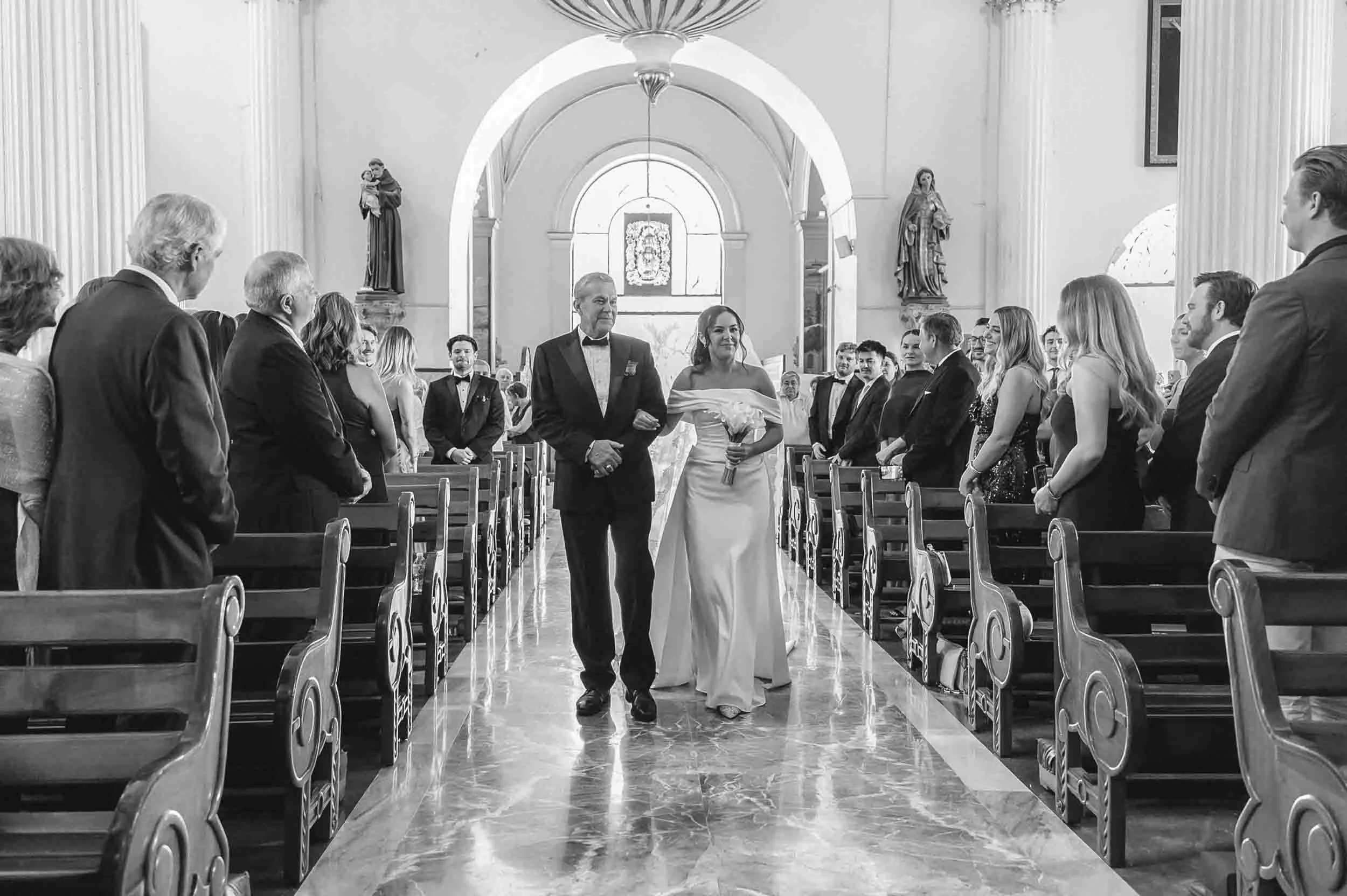 A black-and-white photo of a wedding ceremony inside a church. A bride in a white gown and veil is walking down the aisle escorted by an older man in a tuxedo. Guests are standing on either side of the aisle, watching and smiling.