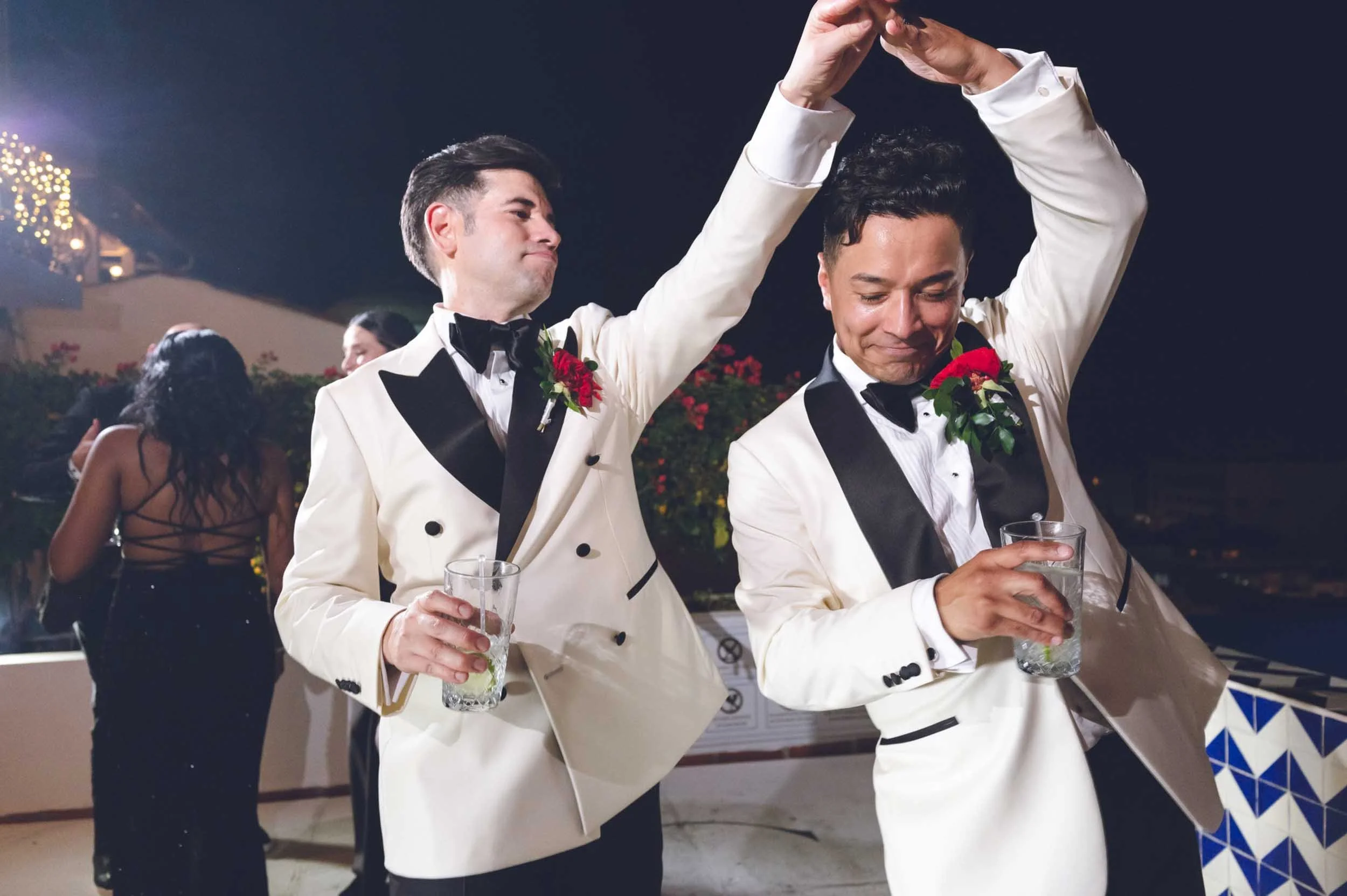 Dancing gay couple in tuxedos with boutonnieres, holding glasses of drinks, at their wedding reception with other guests and decorated surroundings.