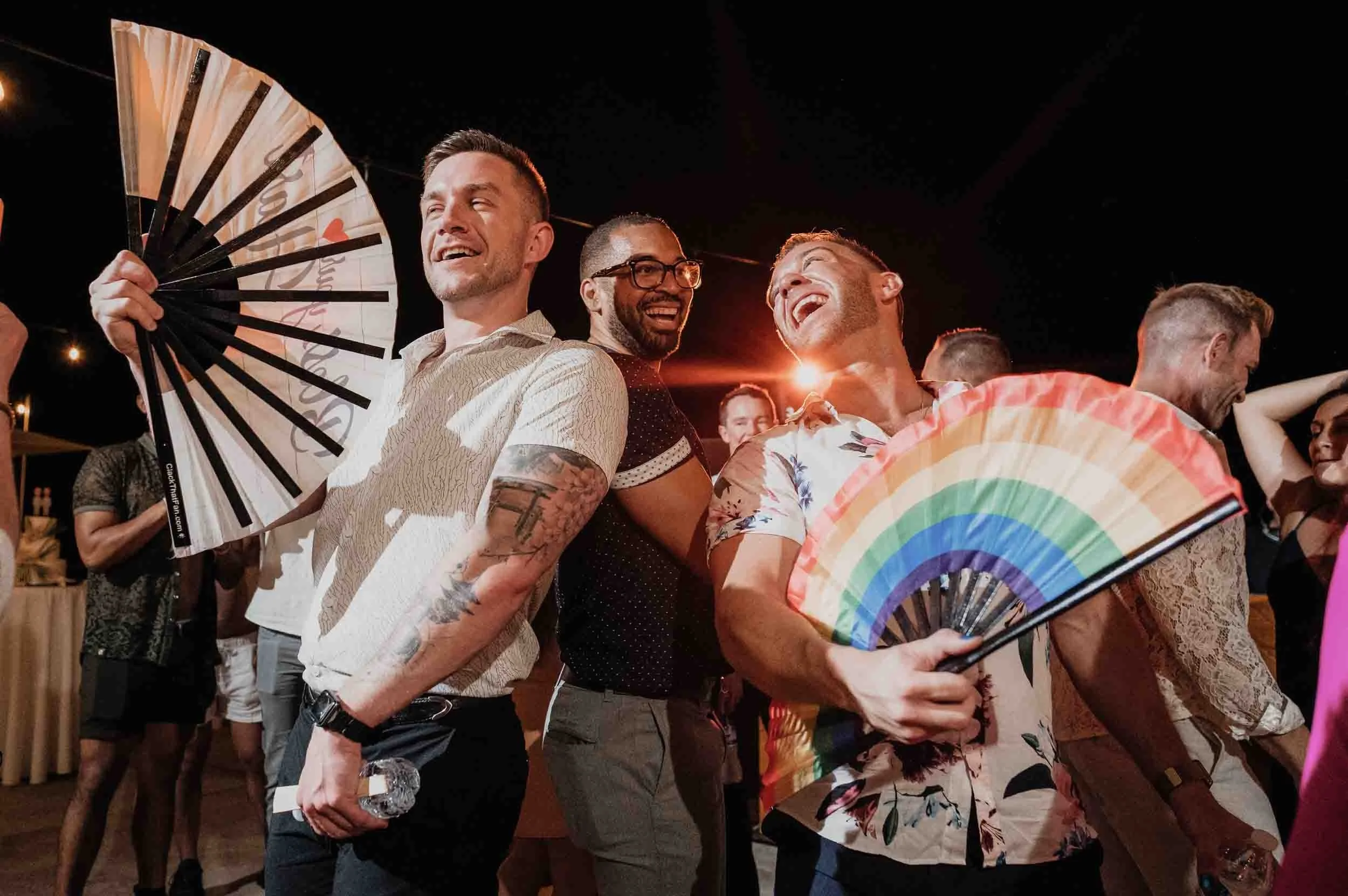 A group of people at a LGBTQ wedding, holding a rainbow fan and smiling with each other.