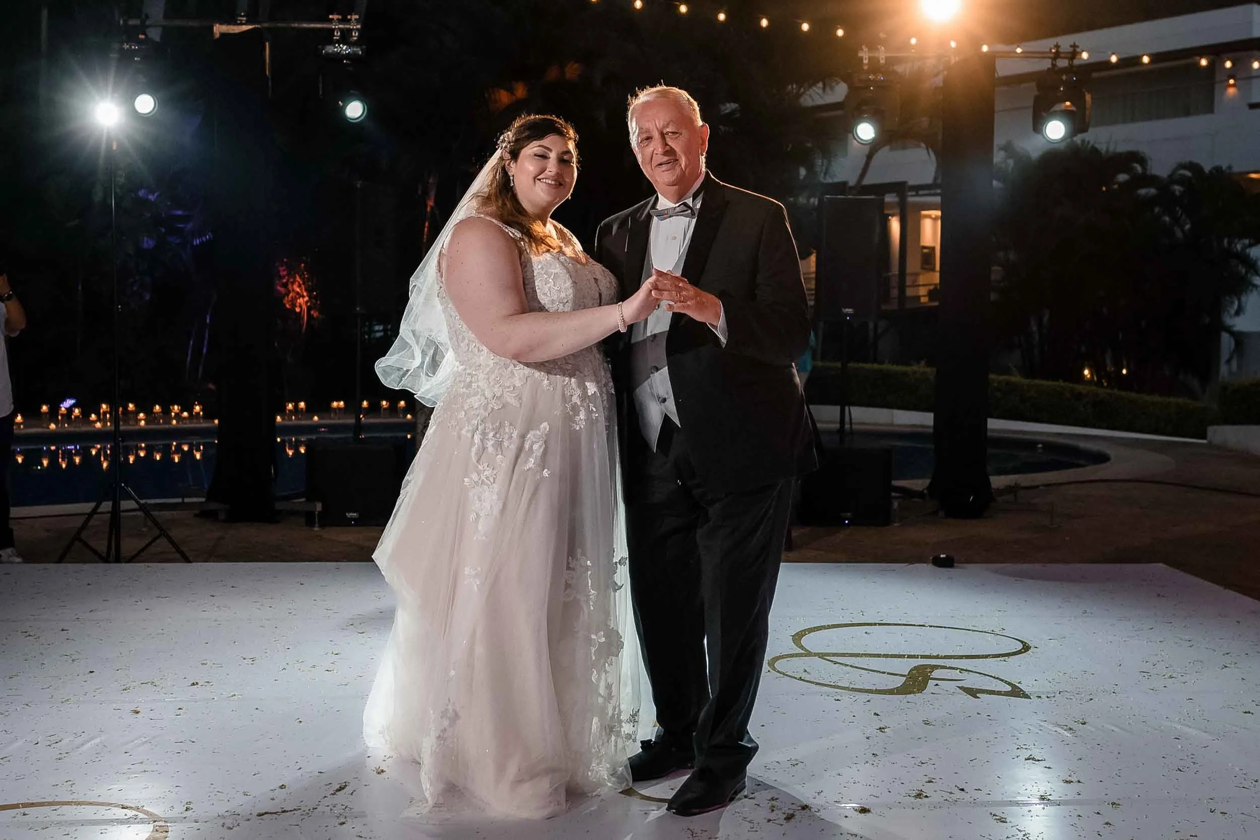 A bride and her father are dancing at a wedding reception outdoors at night.  They are holding hands, smiling, and enjoying the moment, with string lights and a dark background behind them.