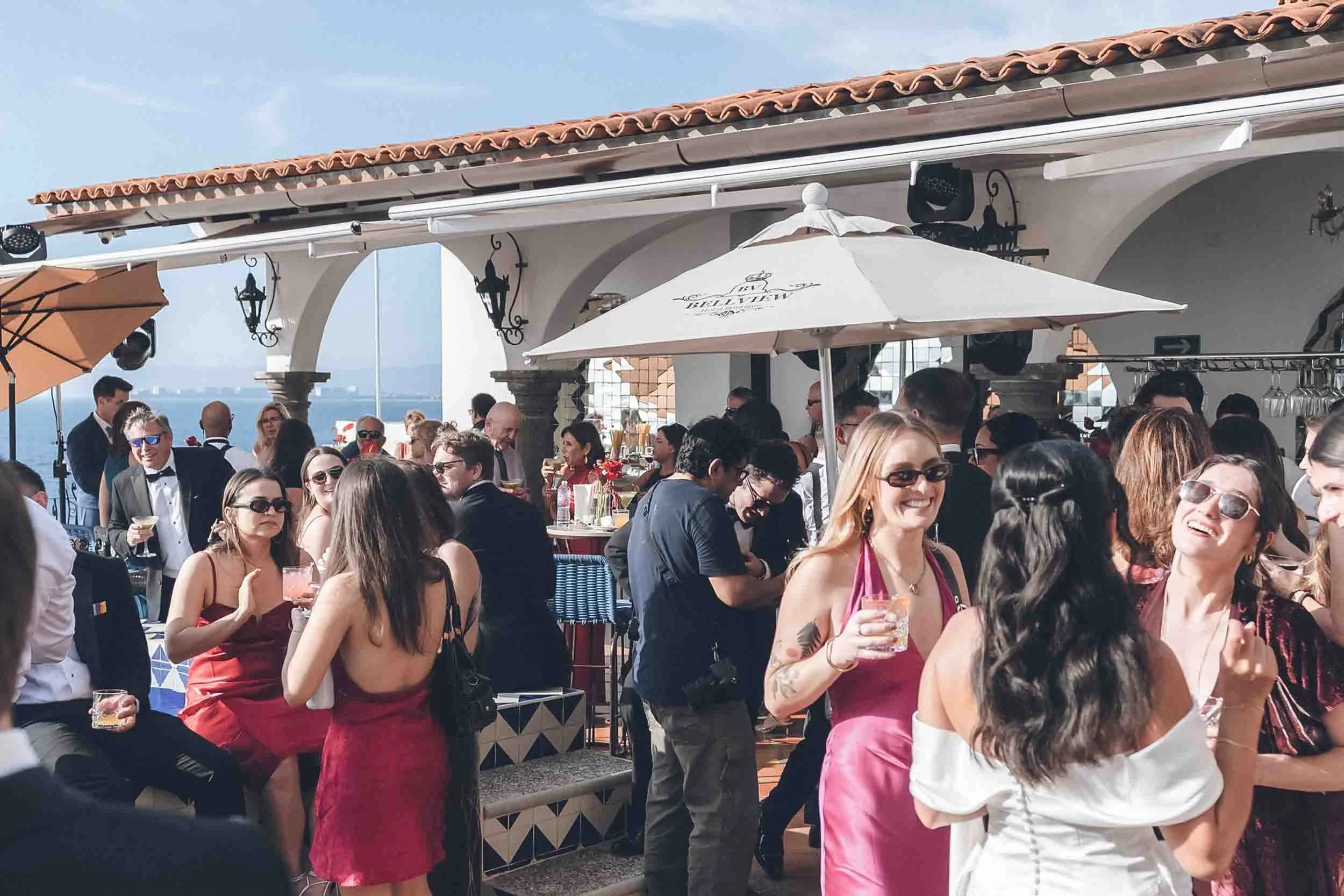 Group of people at an outdoor wedding, some holding drinks, under umbrellas and parasols, with a view of the sea in the background.