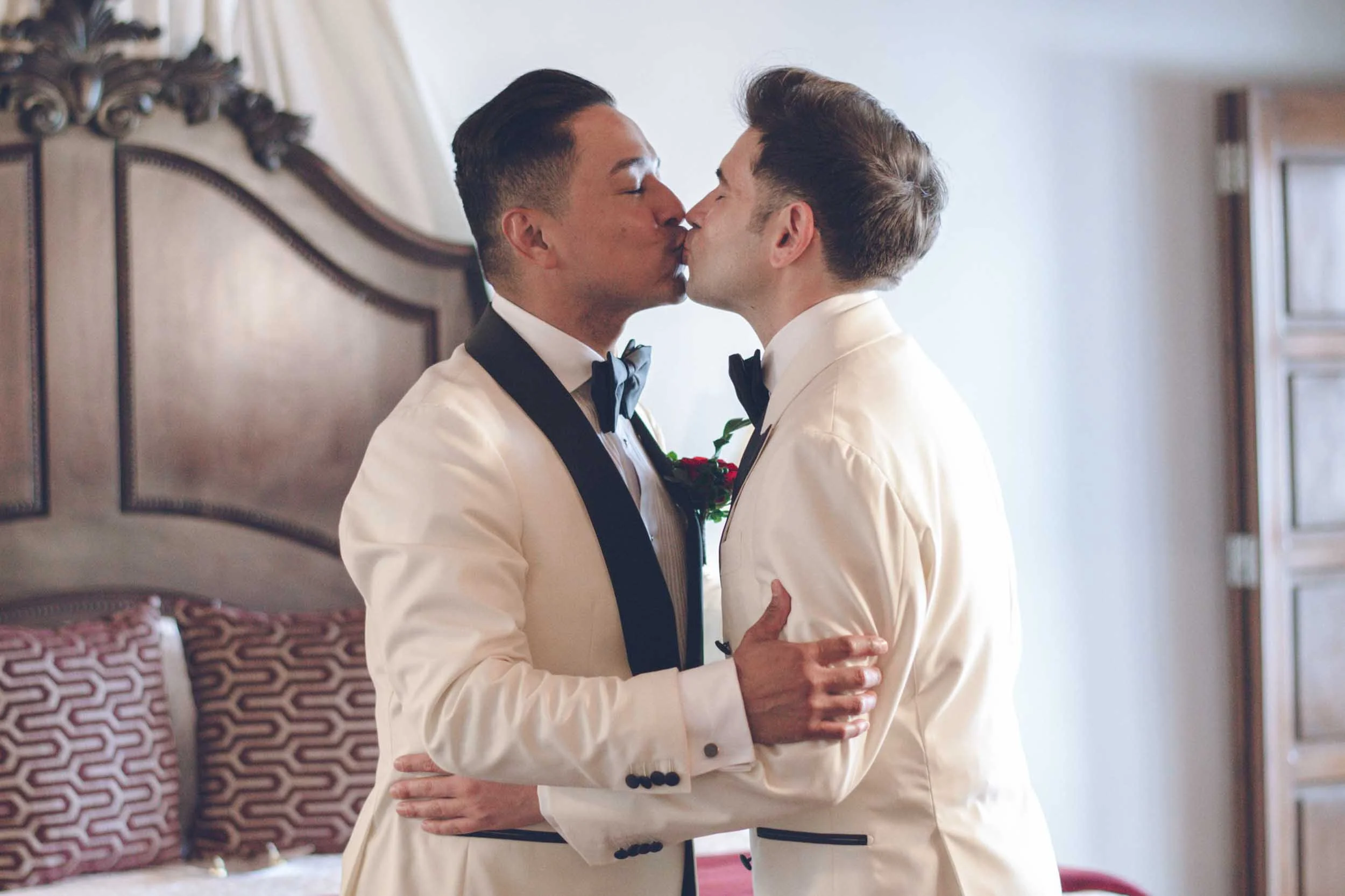 Gay couple in white tuxedos and black bow ties share a kiss  room before their wedding ceremony.
