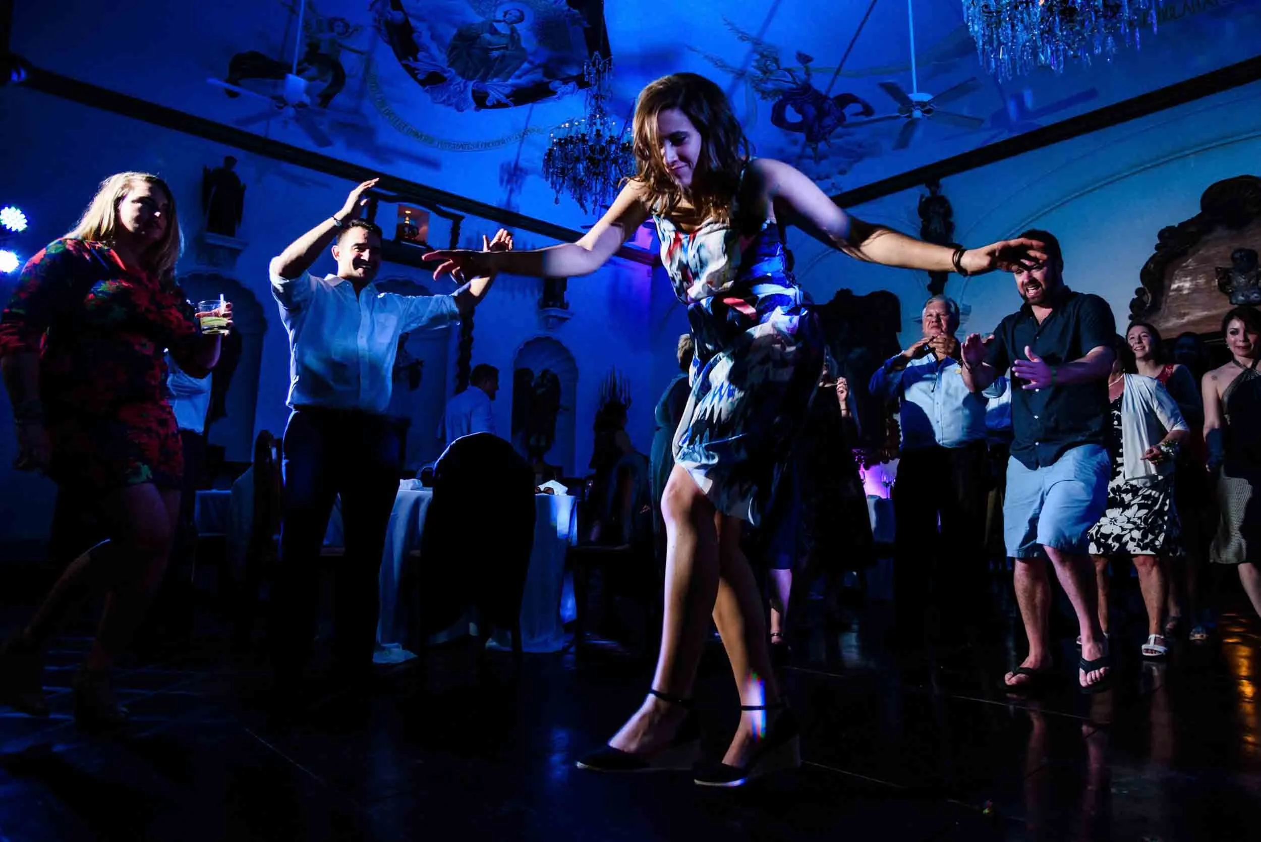 People dancing and celebrating in a dimly lit indoor space with colorful lights, a chandelier, and decorative ceiling art.