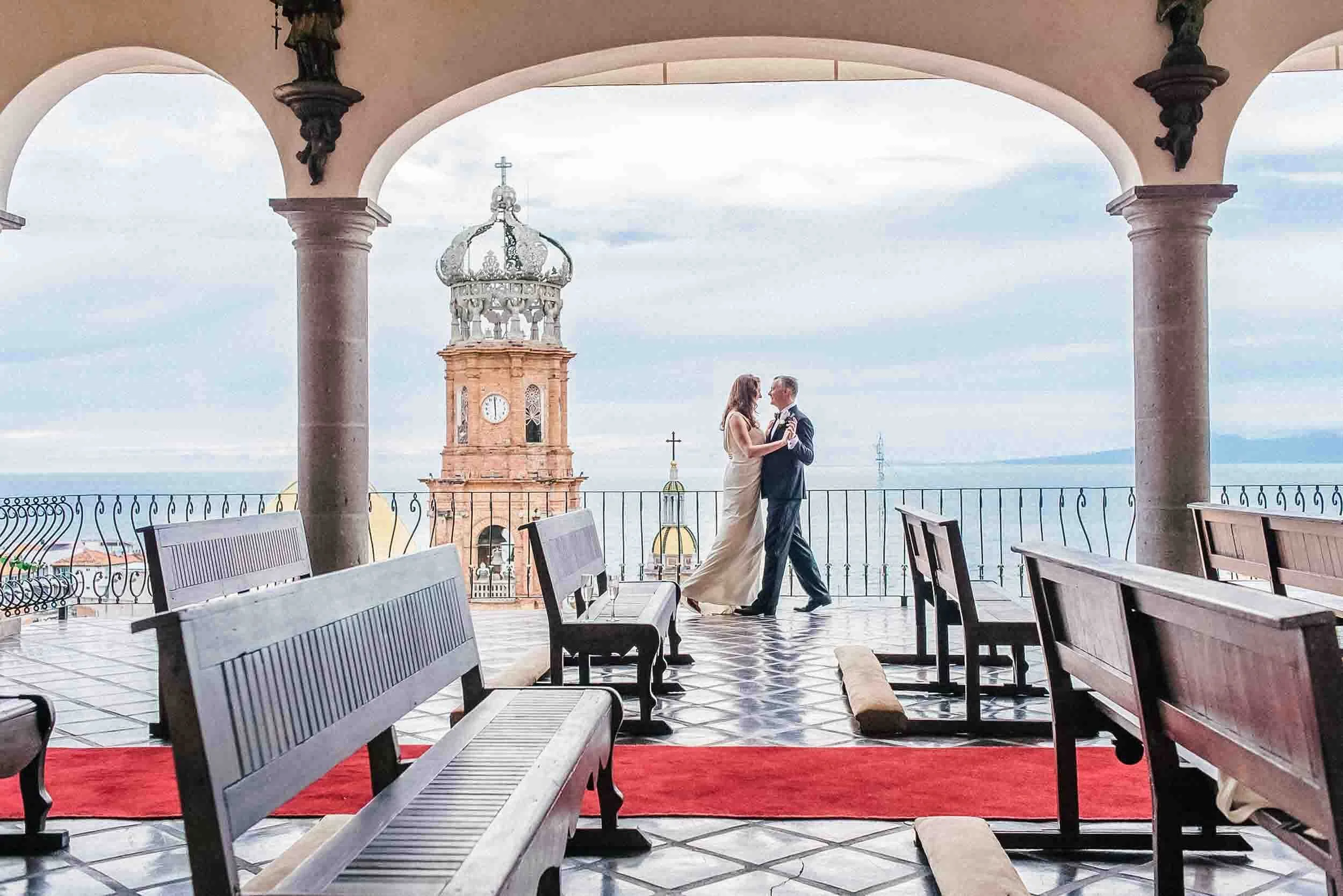 Bride and groom dancing on a balcony with a view of a Puerto Vallarta church with the clock tower and the sea in the background, under an archway with statues.