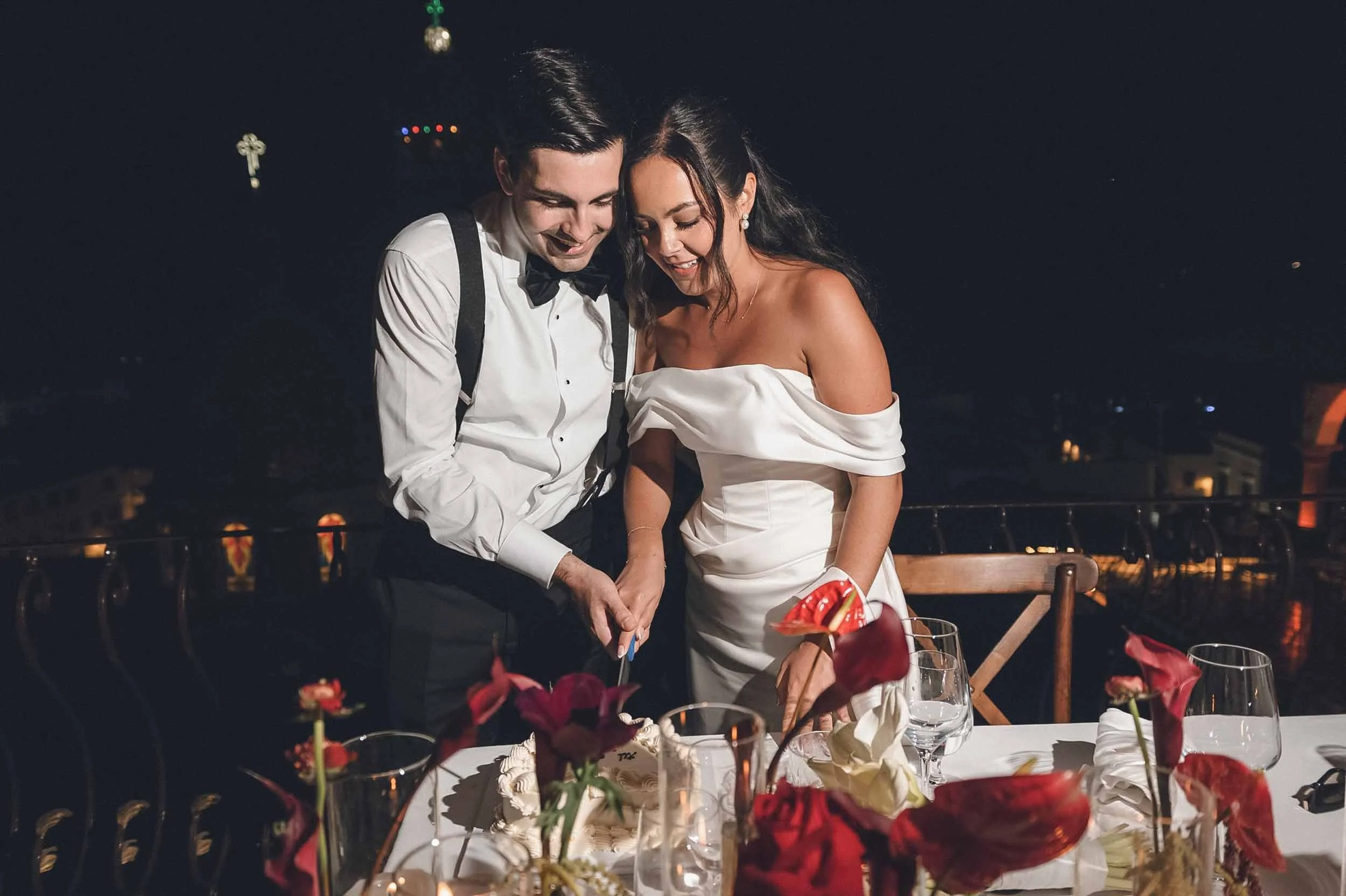 Bride and groom smiling during cake cutting  at their wedding reception.The man is wearing a tuxedo with a bow tie, and the woman is in a white off-shoulder wedding dress. 