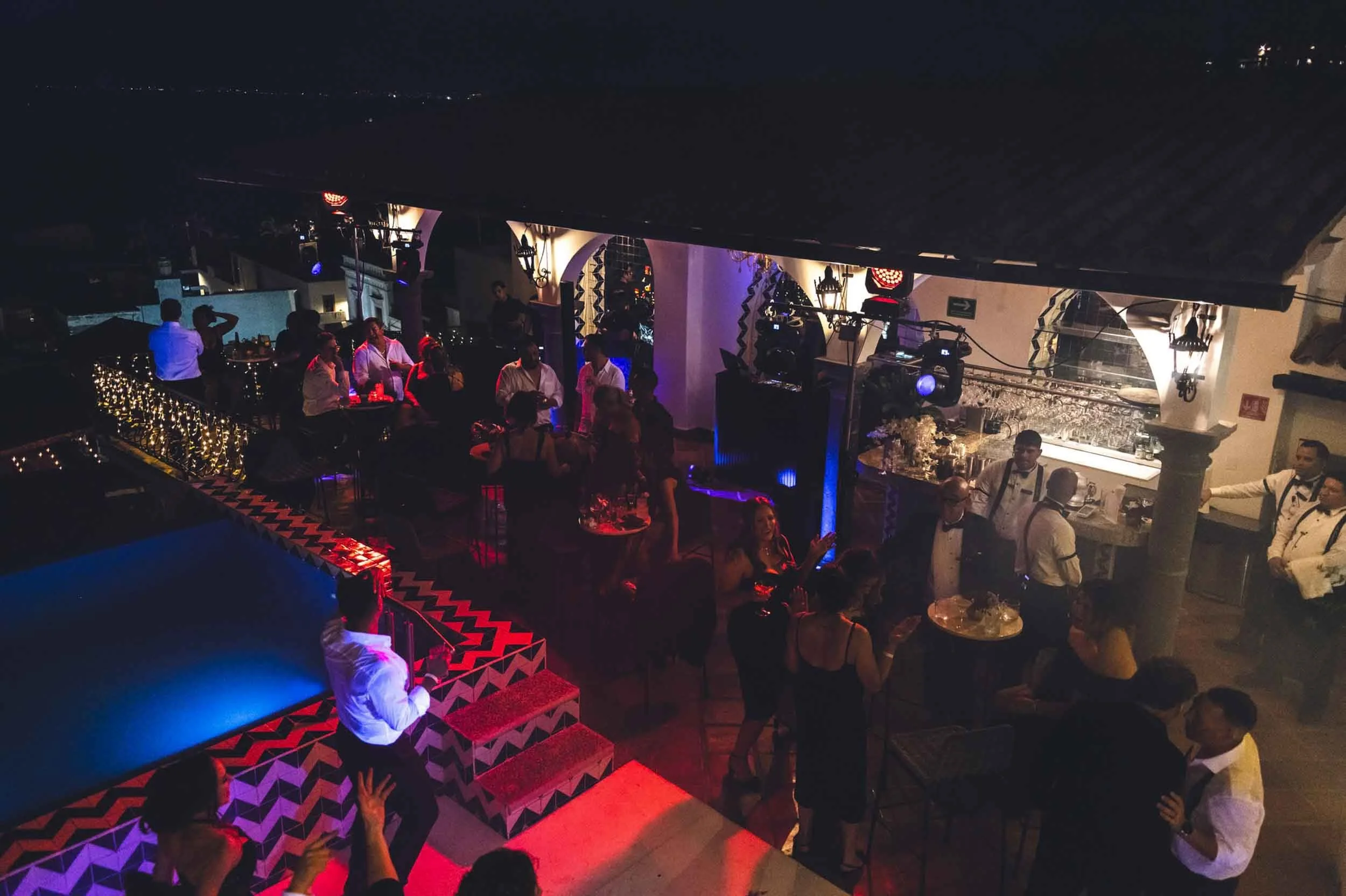 People socializing and dancing at wedding reception with colorful lighting, bar area, and decorative architectural elements.