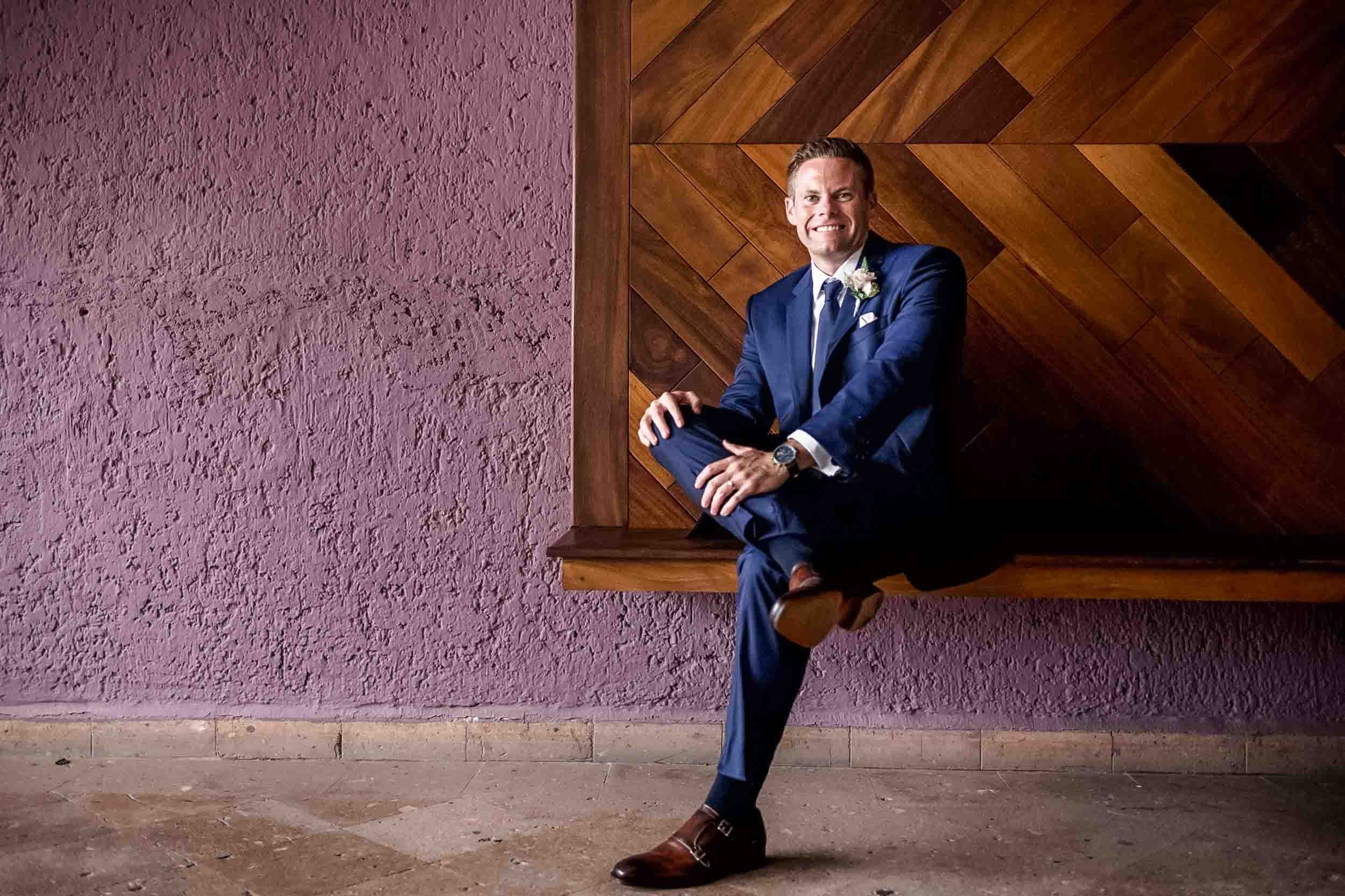 Groom in a dark blue suit sits on a floating wooden ledge against a textured pink wall with a wood-paneled section behind him. 