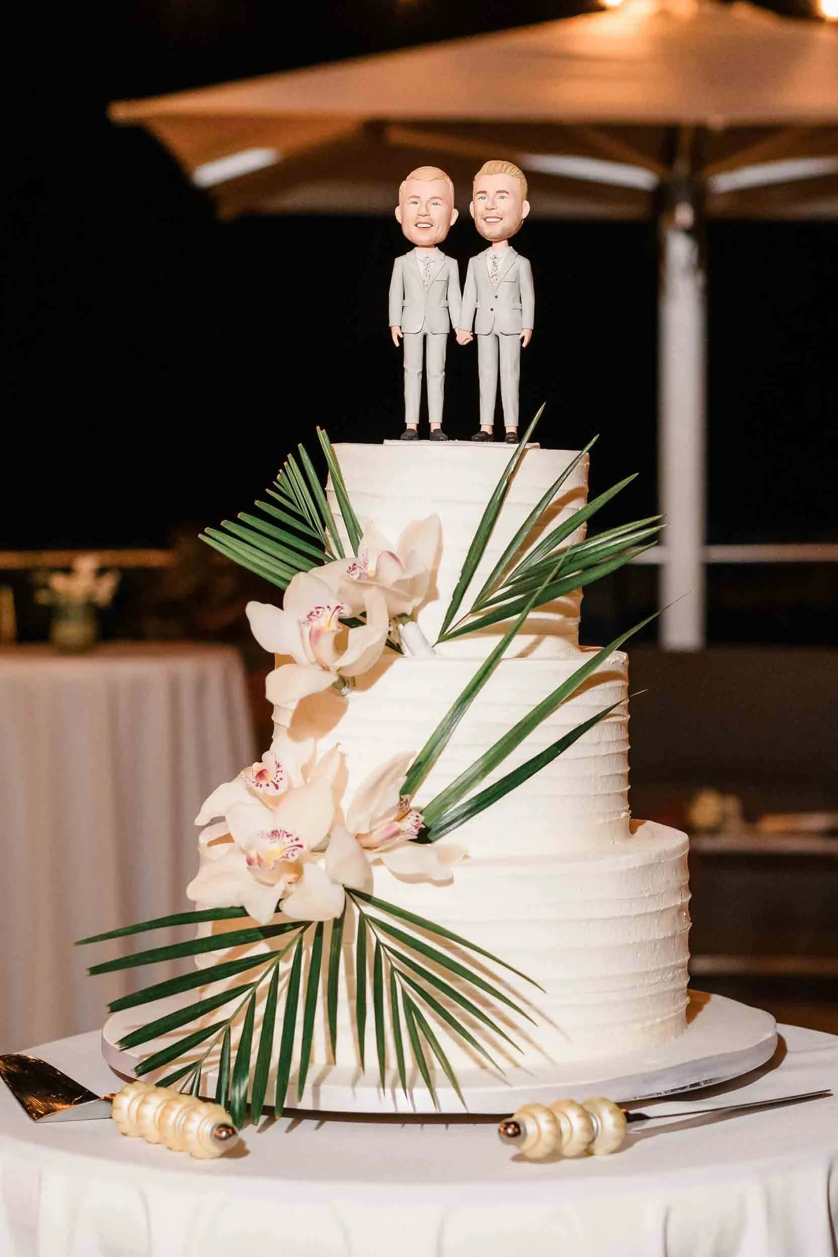 A wedding cake with two groom figurines on top, decorated with white frosting, orchids, and palm leaves.