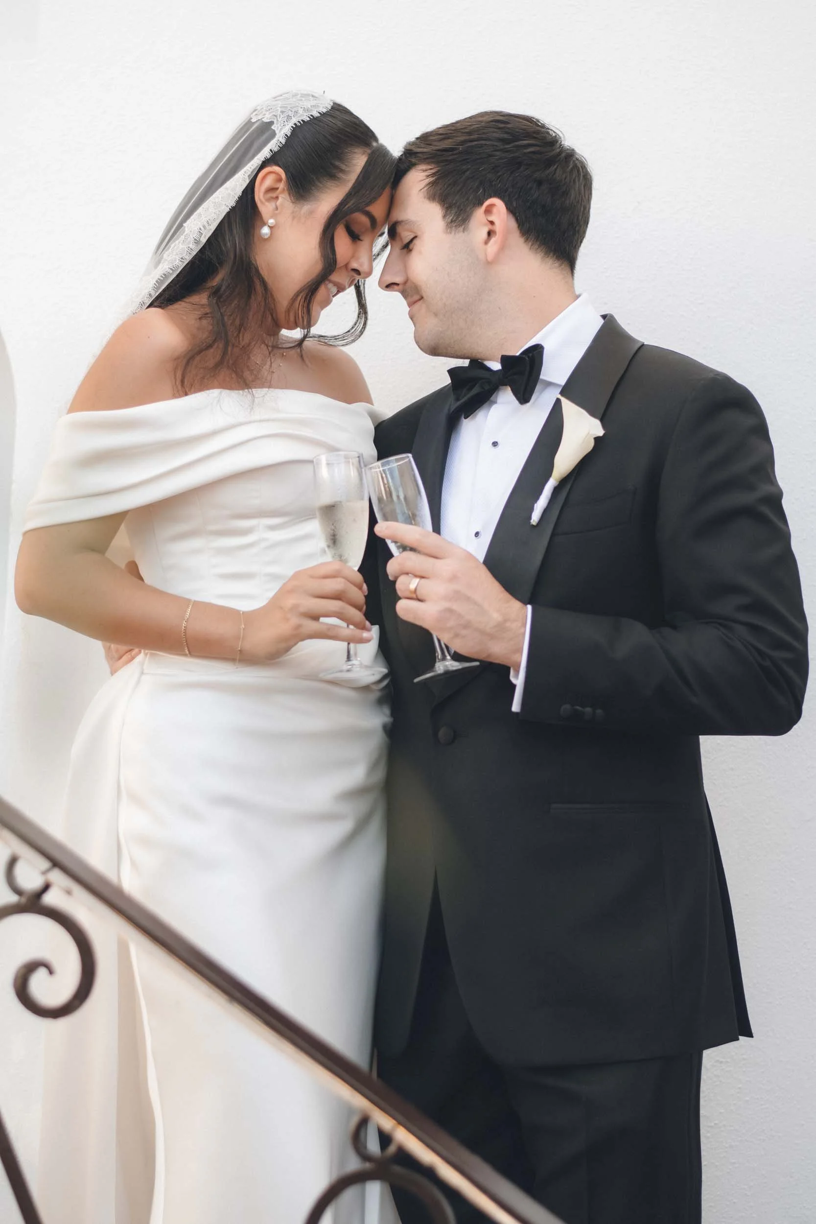A newlywed couple, dressed in wedding attire, are holding champagne glasses while touching foreheads and smiling, standing against a plain white wall.