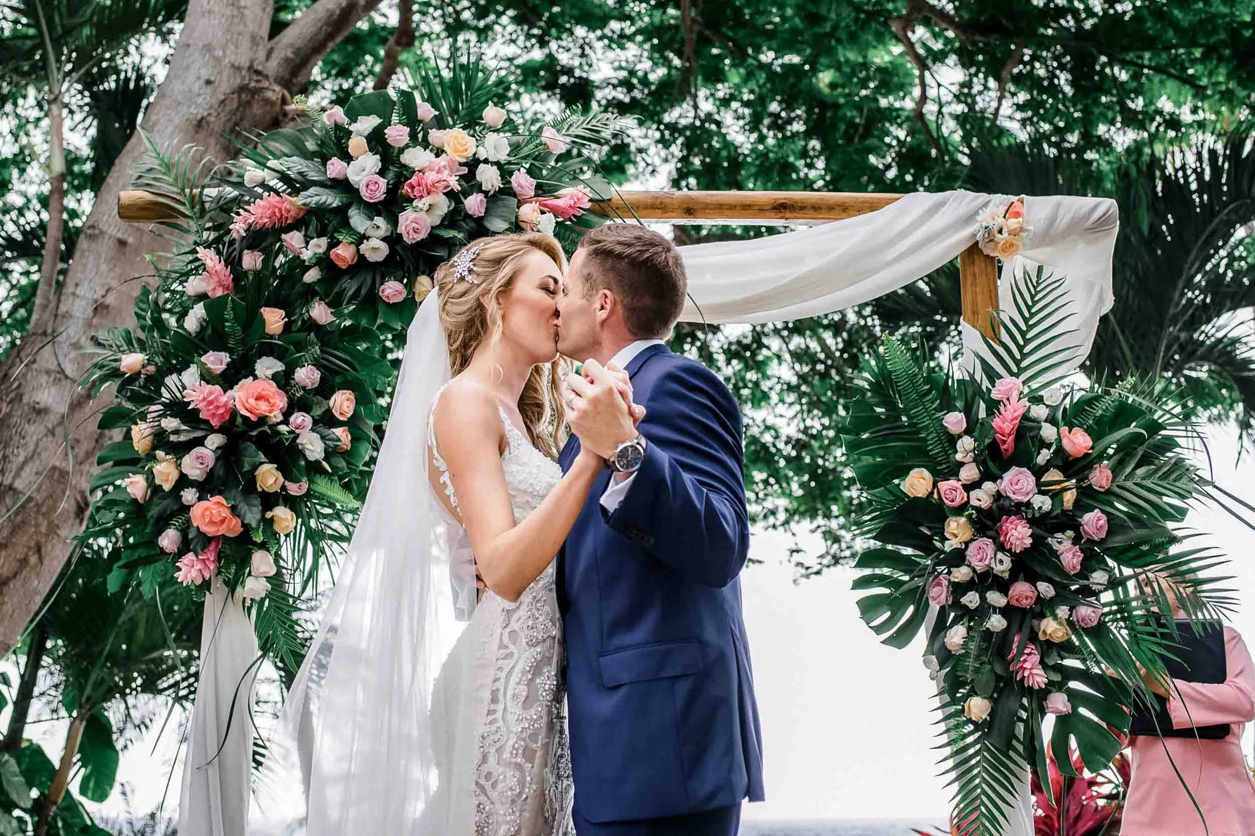 A bride and groom kiss during their outdoor wedding ceremony under a decorated arch of flowers and greenery.