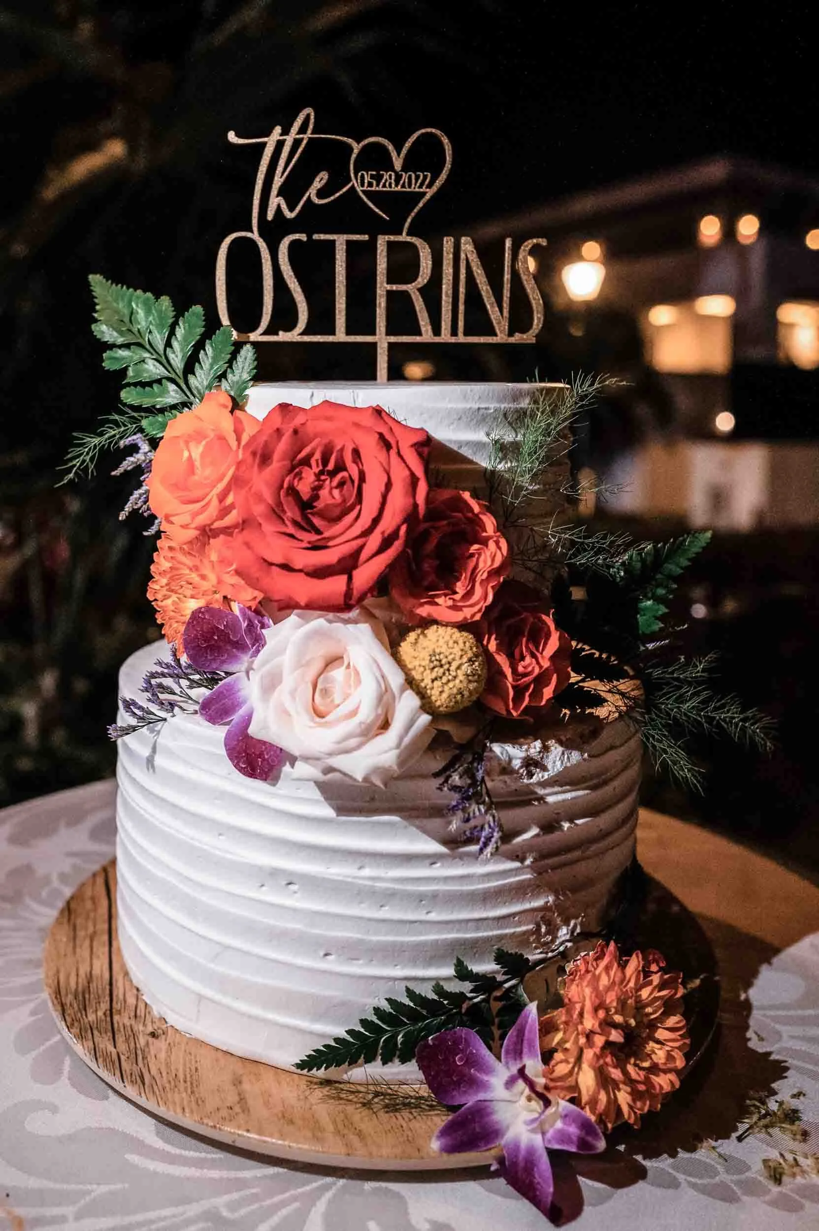 A wedding cake decorated with fresh flowers, including roses and orchids, on a wooden cake stand. A topper with the inscription "the Ositrins" and a heart with the date 05.28.2022 is placed on the cake. The background is dimly lit with warm lights.