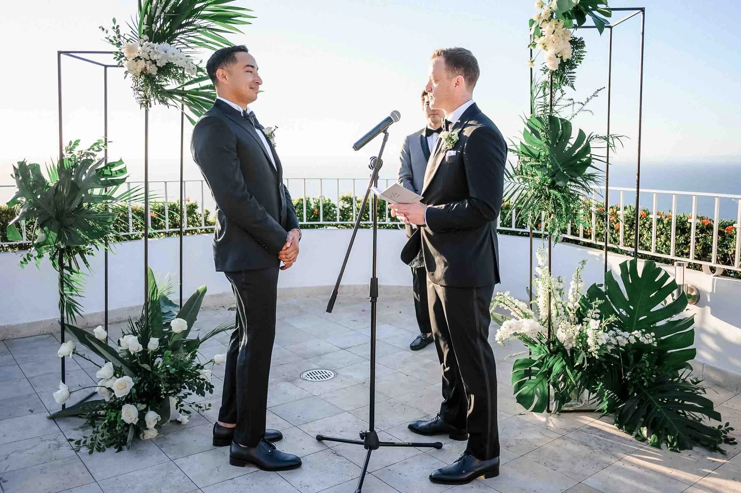 Gay couple in tuxedos,, facing each other during their wedding ceremony with a officiant in the background, surrounded by green tropical leaves and white flowers, on a terrace with ocean view.