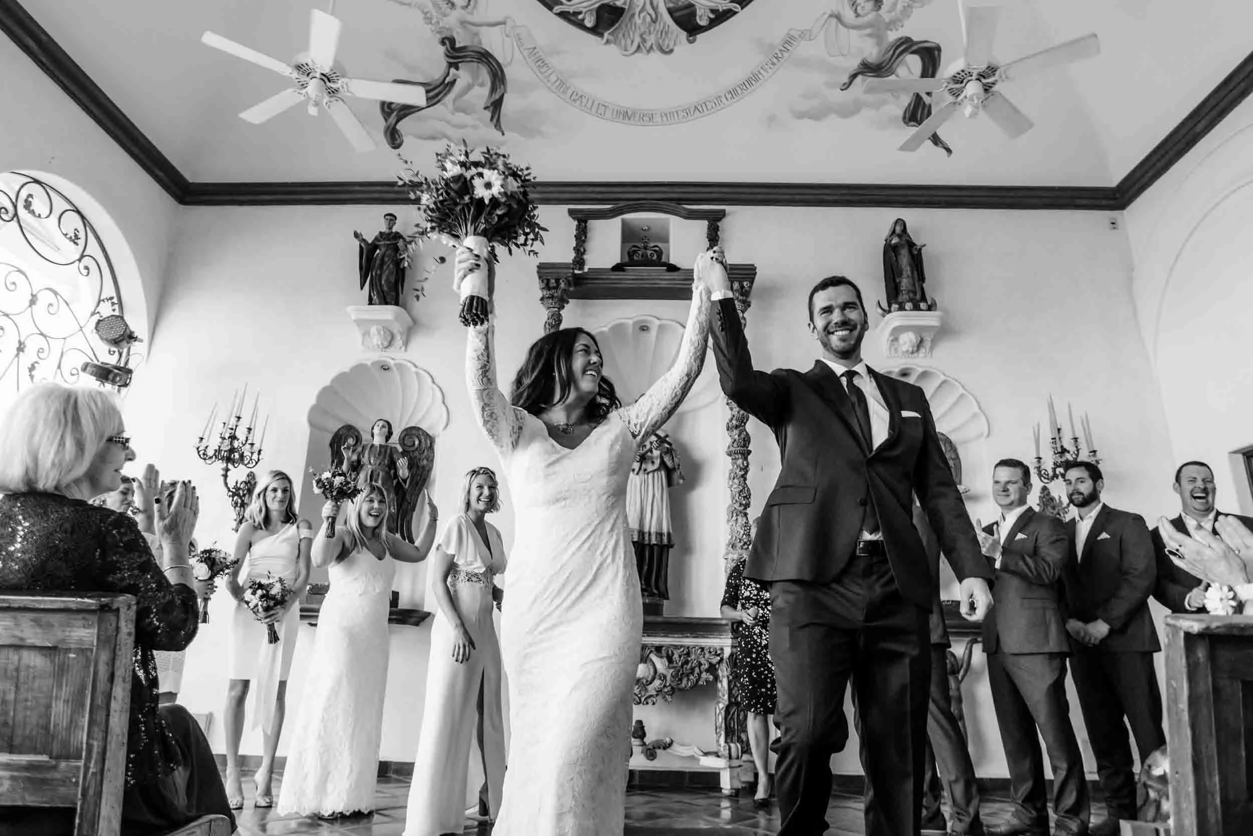 A newly married couple holding hands up in the air and smiling exiting their wedding ceremony surrounded by friends and family, with religious statues and ornate decorations in the background.
