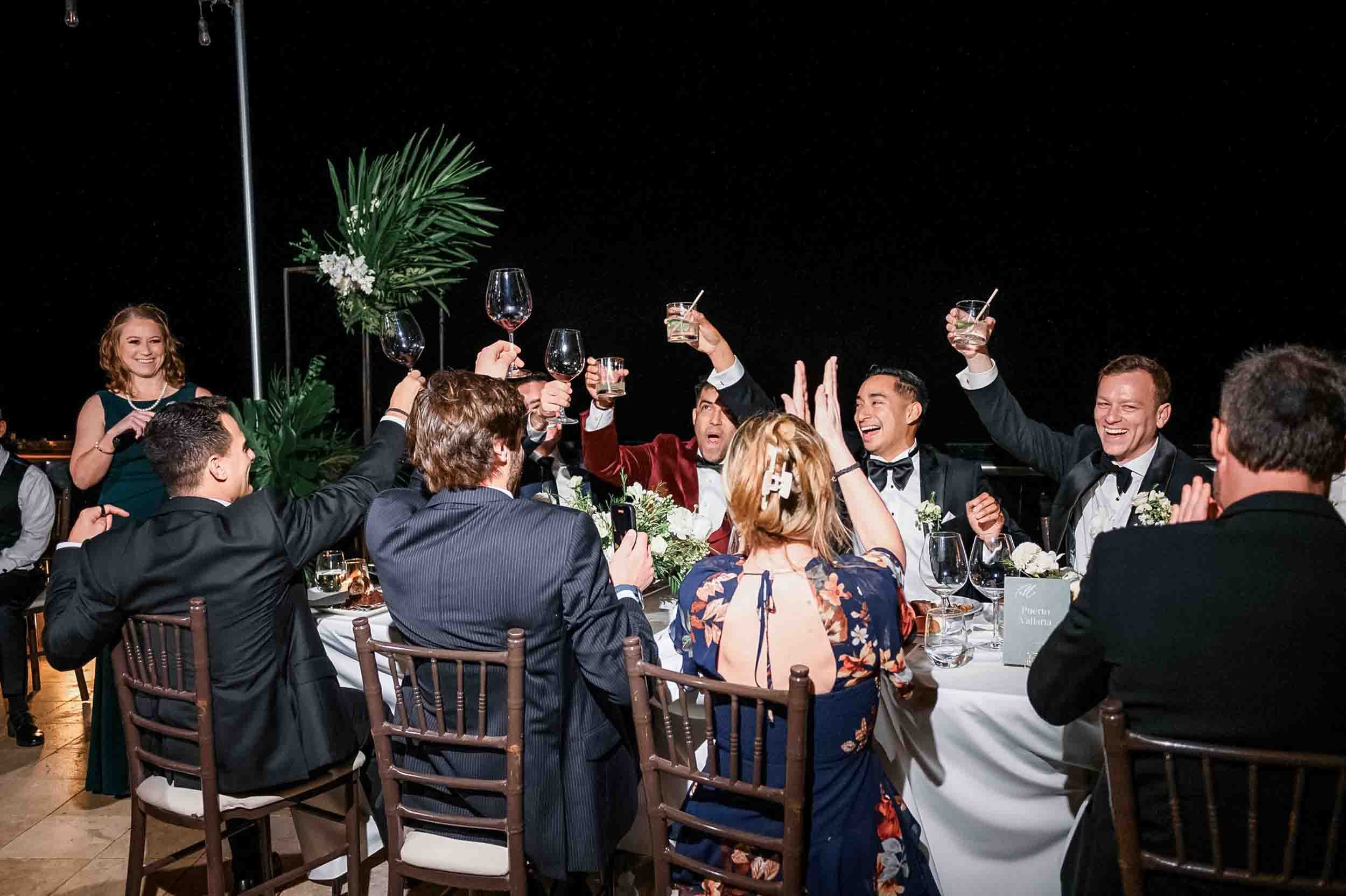 Group of people celebrating and toasting at a wedding reception, with smiling faces, a woman in a green dress standing, dinner table with flowers and glasses, night outdoor setting.