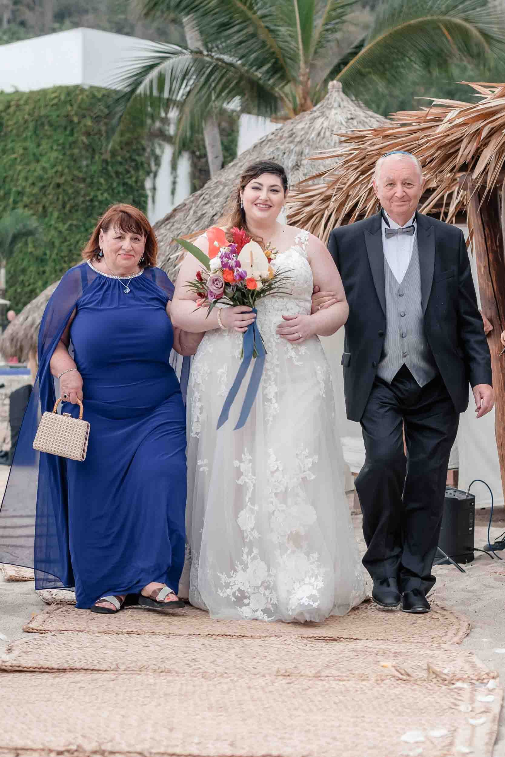 Parents of the bride walking her down the asile, on a beach with palm trees and thatched umbrellas.