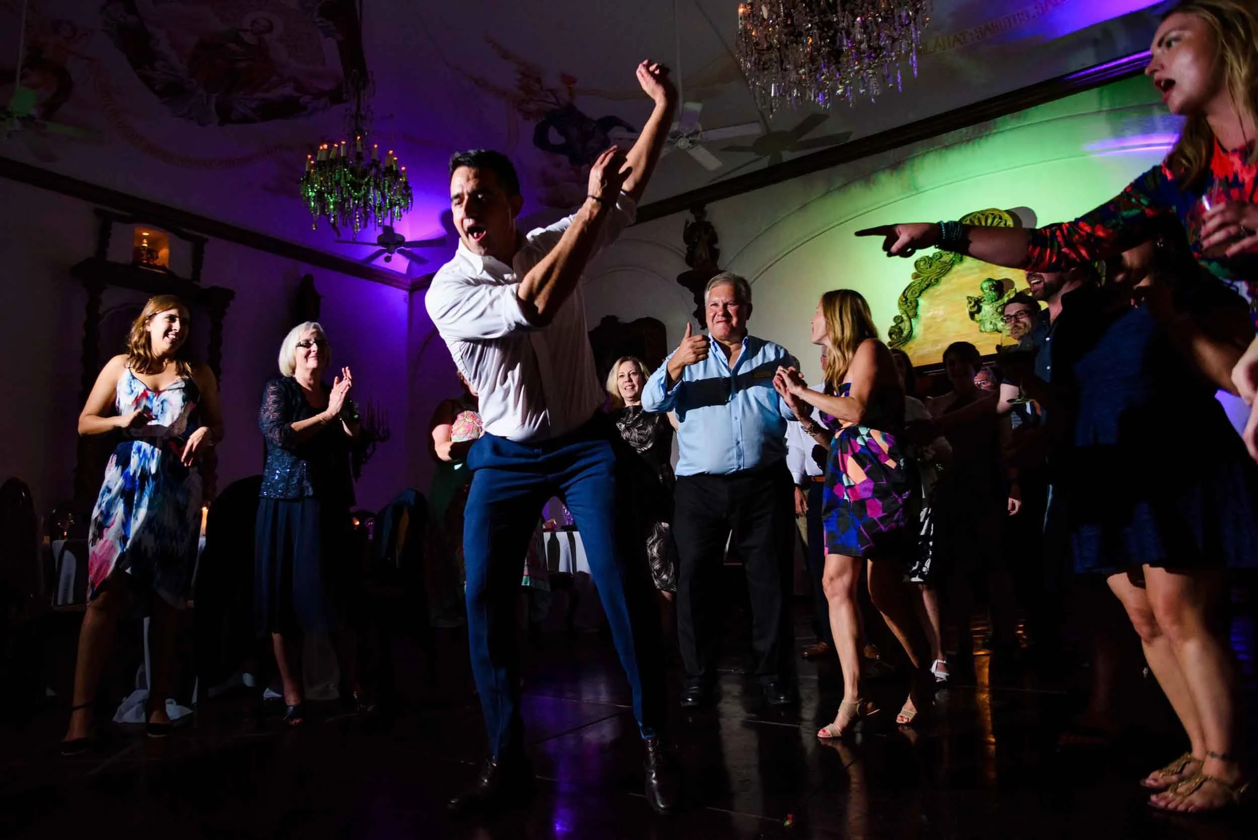 People dancing and having fun at a wedding reception, with colorful lighting and a chandelier overhead.