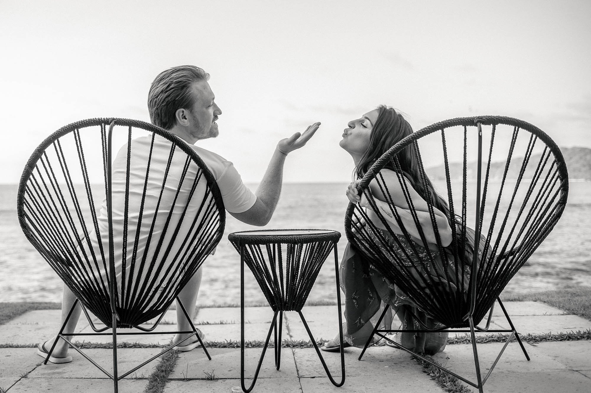 Engagement session, the groom to be is blowing a kiss to his fiancé, both are sitting down in Acapulco chairs.