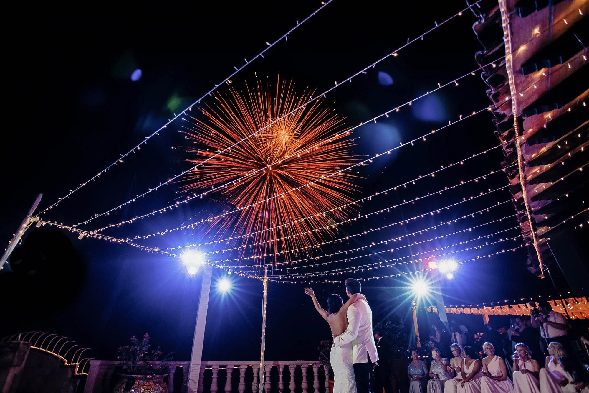 Night scene at a wedding with fireworks in the sky, string lights overhead, and the bride and groom.