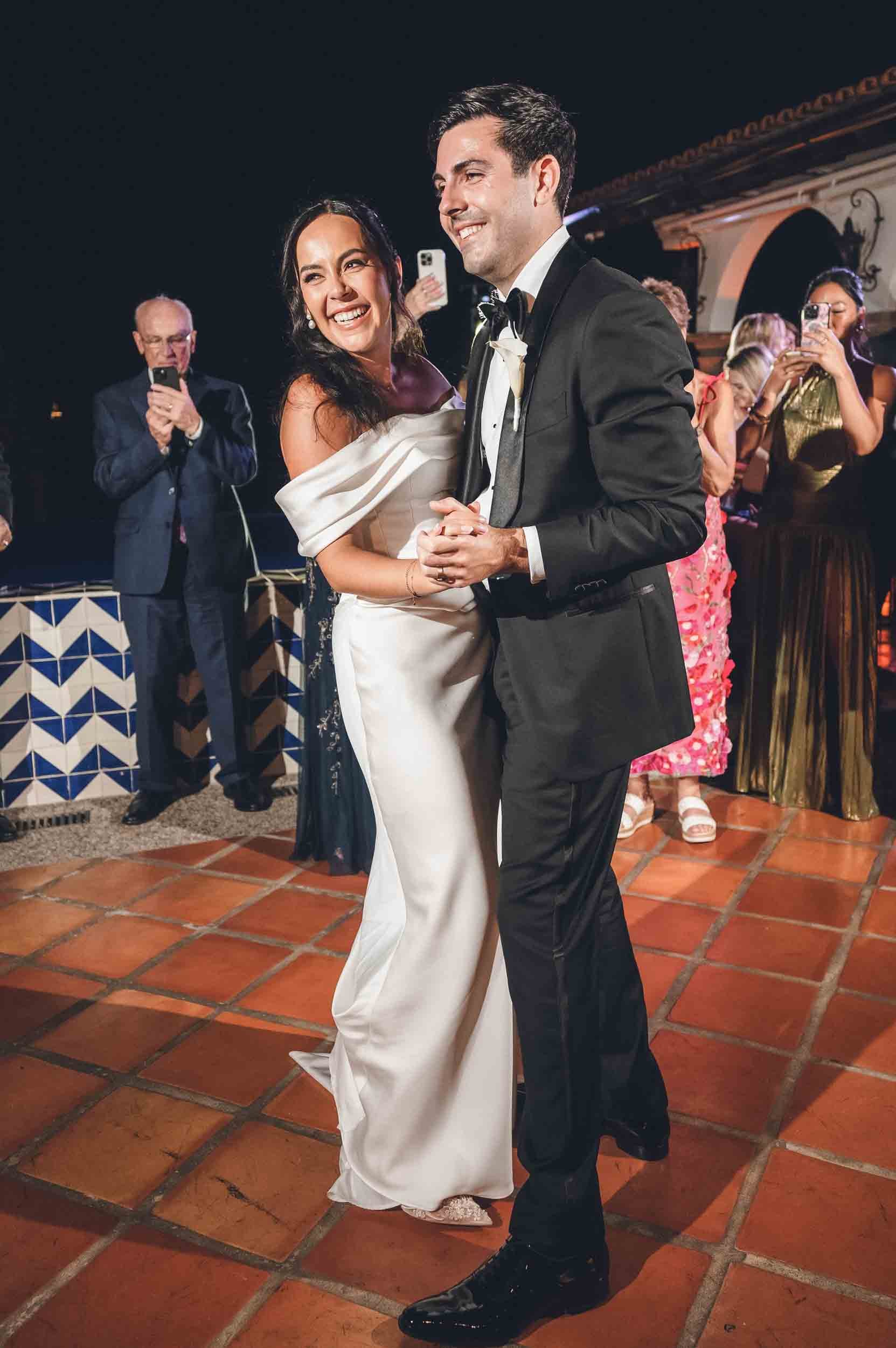 A bride and groom are dancing at their outdoor wedding reception, smiling and holding hands. The bride is wearing a white off-shoulder gown, and the groom is dressed in a black tuxedo. Guests are watching and taking photos in the background.