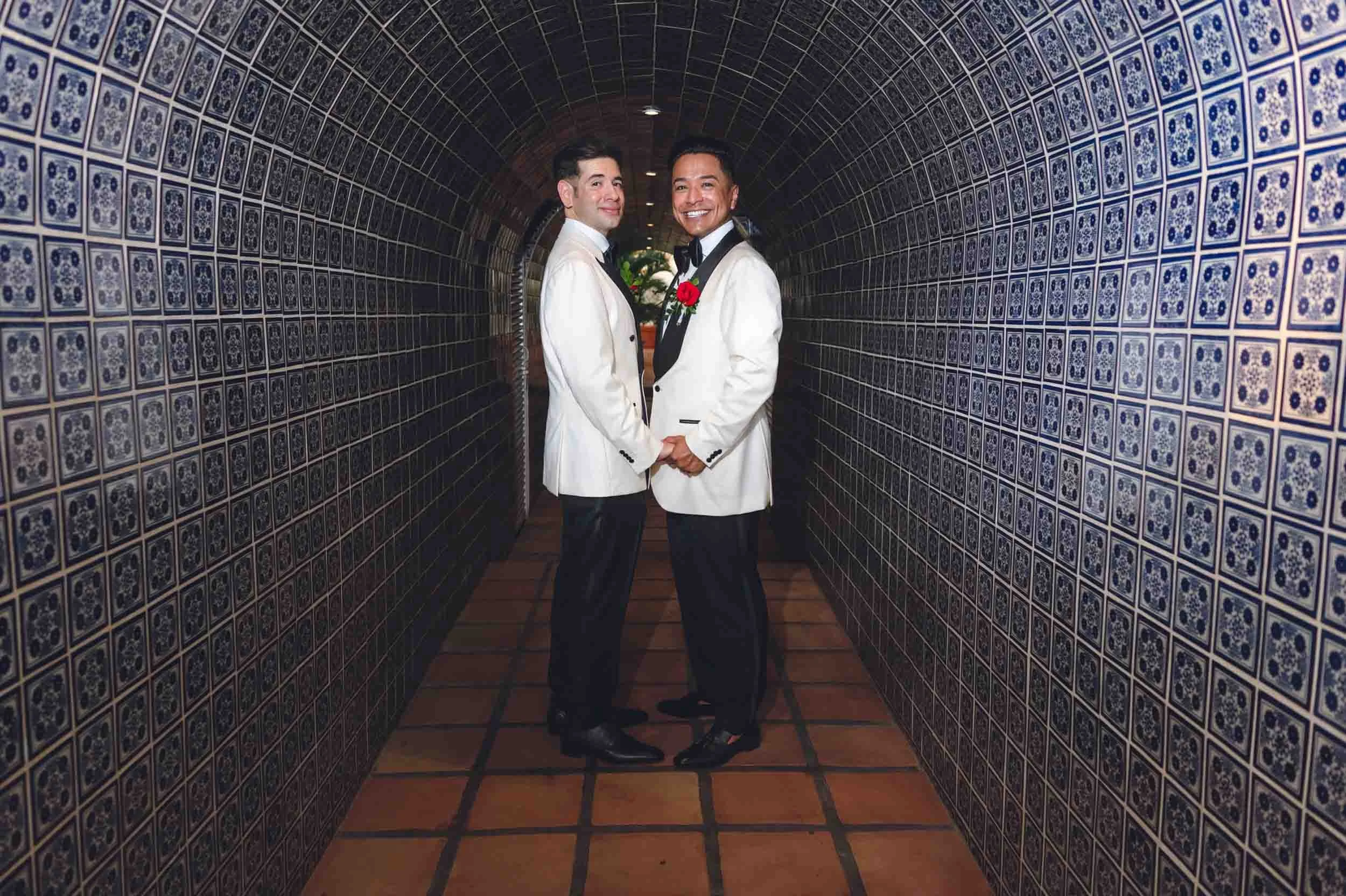 Gay couple dressed in white tuxedos with black bow ties and boutonnières standing in a narrow corridor lined with decorated tiles on the walls.