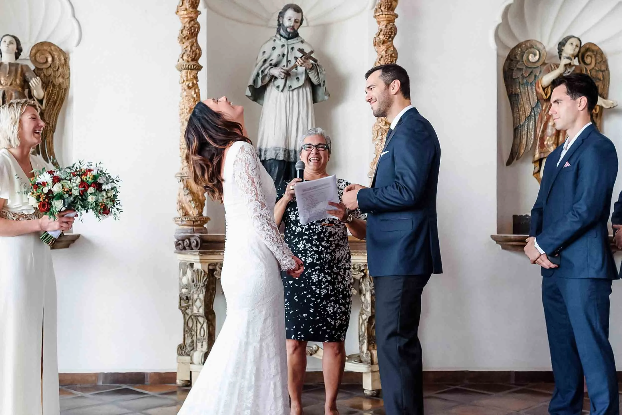 A wedding ceremony  at La Capella in Puerto Vallarta with a bride and groom standing face-to-face, holding hands, surrounded by an officiant and other guests. there are religious statues and intricate wooden carvings in the background.