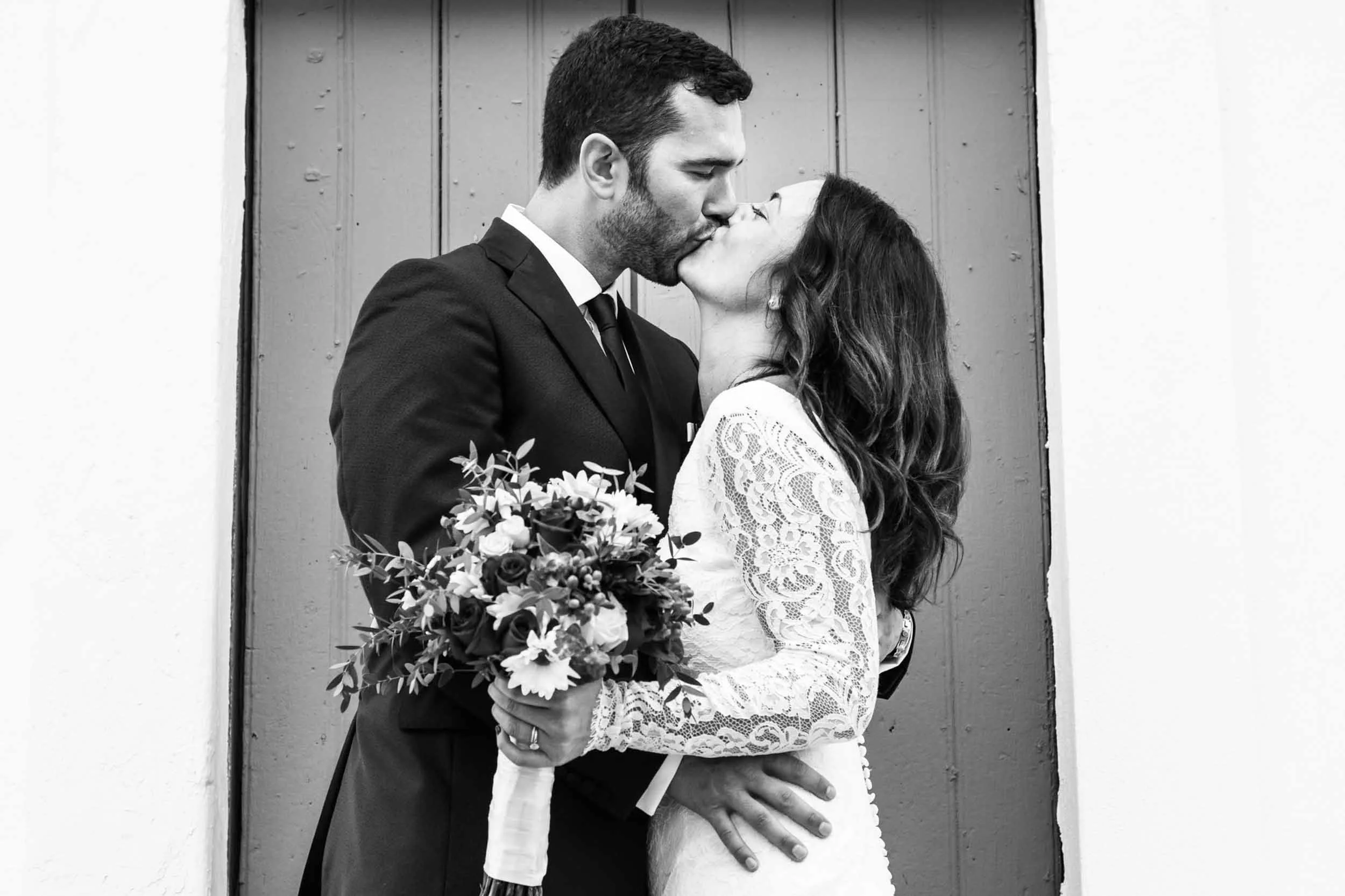 A black and white photo of a couple kissing, dressed in wedding attire, with the groom in a suit and the bride in a lace dress holding a bouquet of flowers.