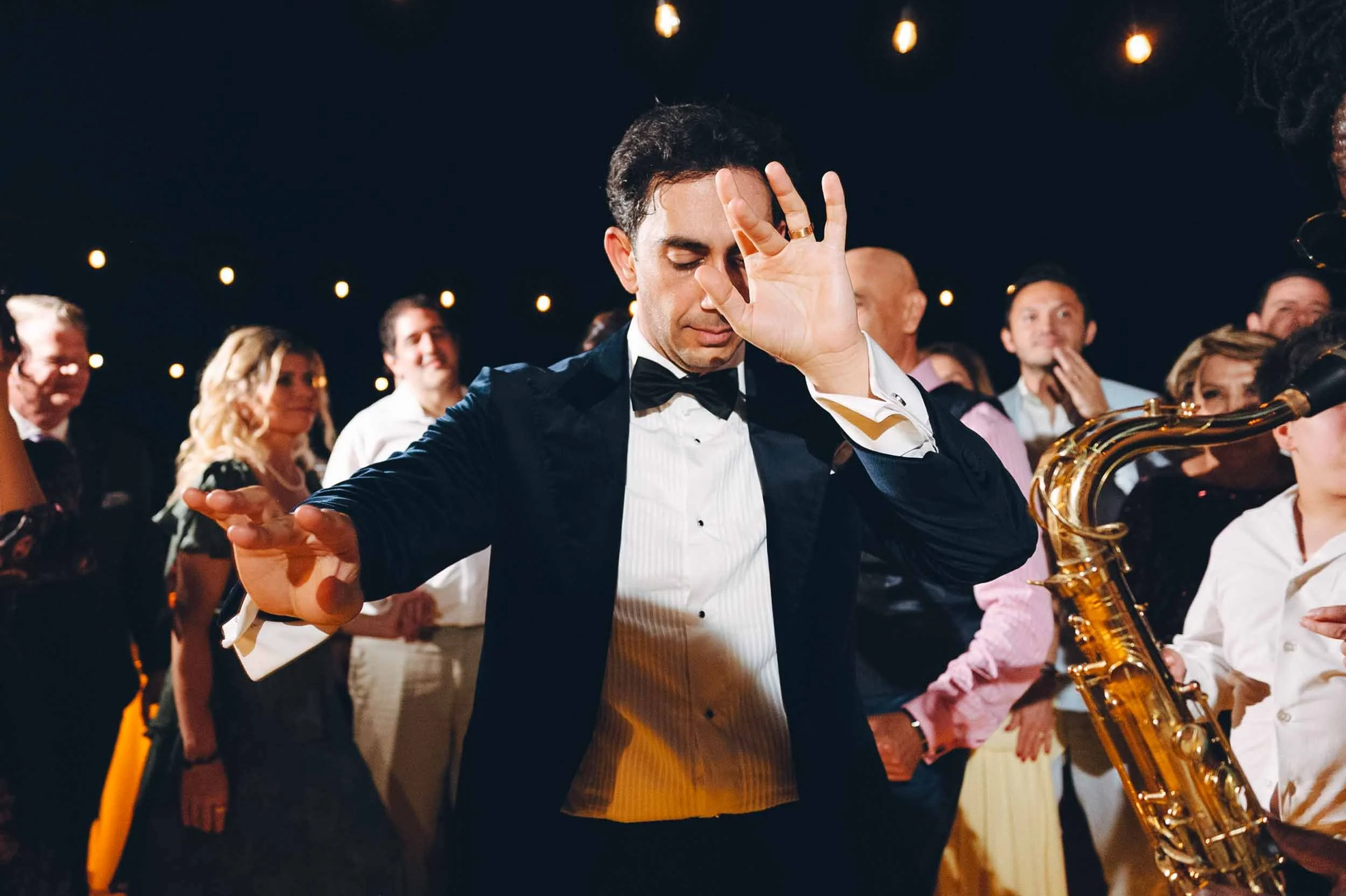 Avgroom in a tuxedo and bow tie dancing on his wedding celebration, surrounded by other people, some watching and smiling.