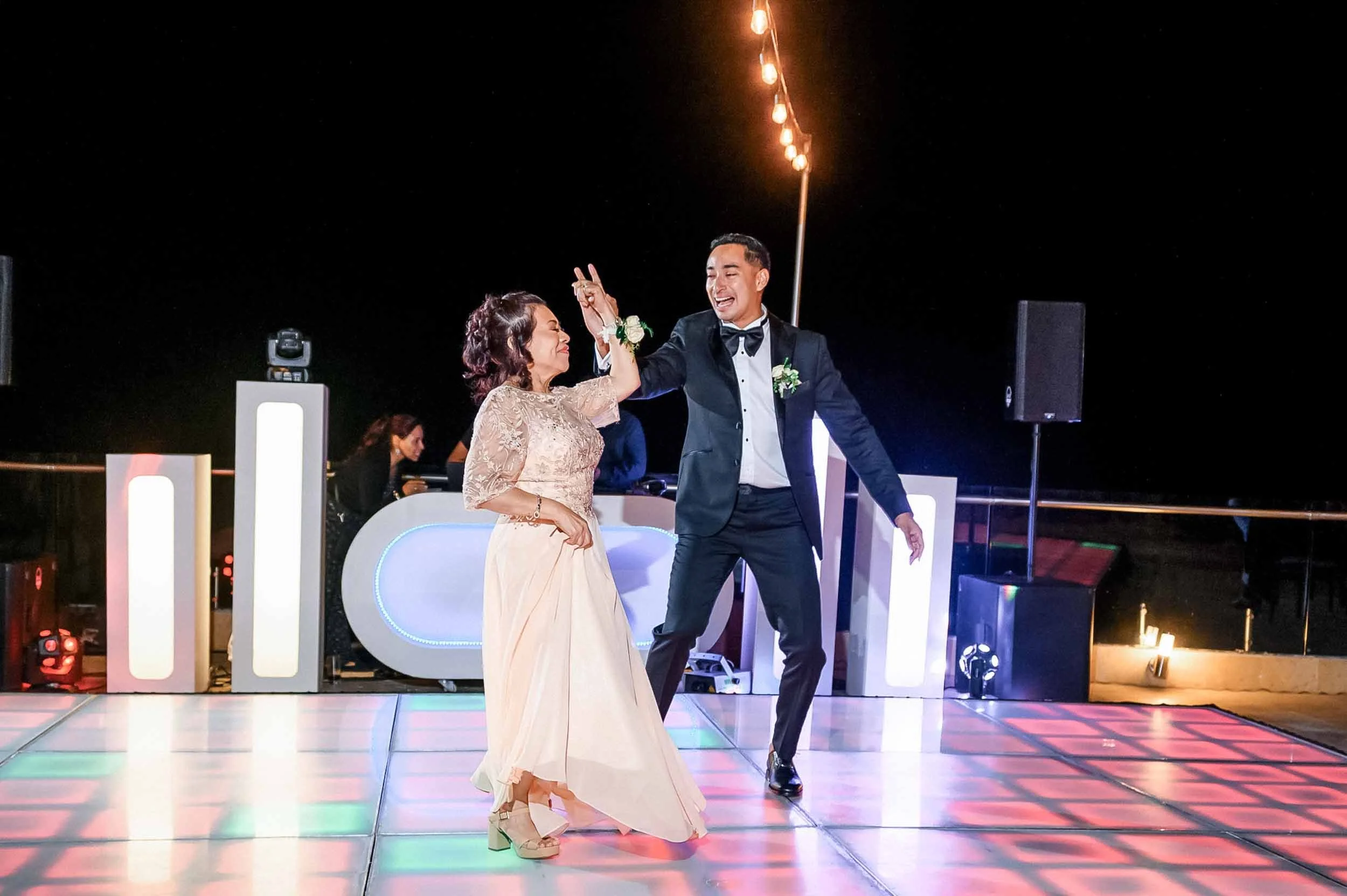 Groom and his mother dancing during the wedding reception under string lights at night. 
