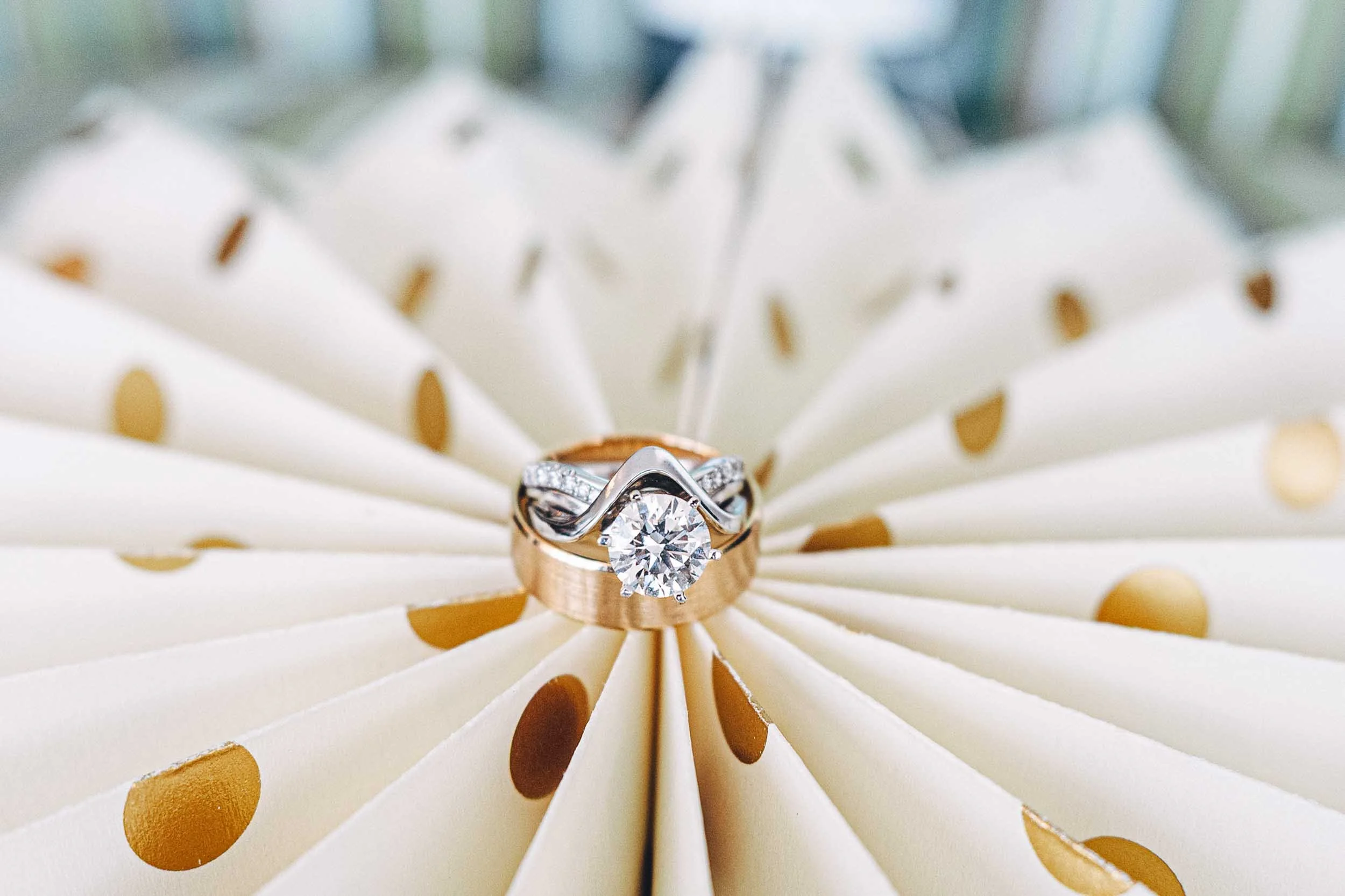A diamond engagement ring with a round cut diamond set in a silver prong setting, placed on a gold band, resting on a white and gold polka-dotted surface.