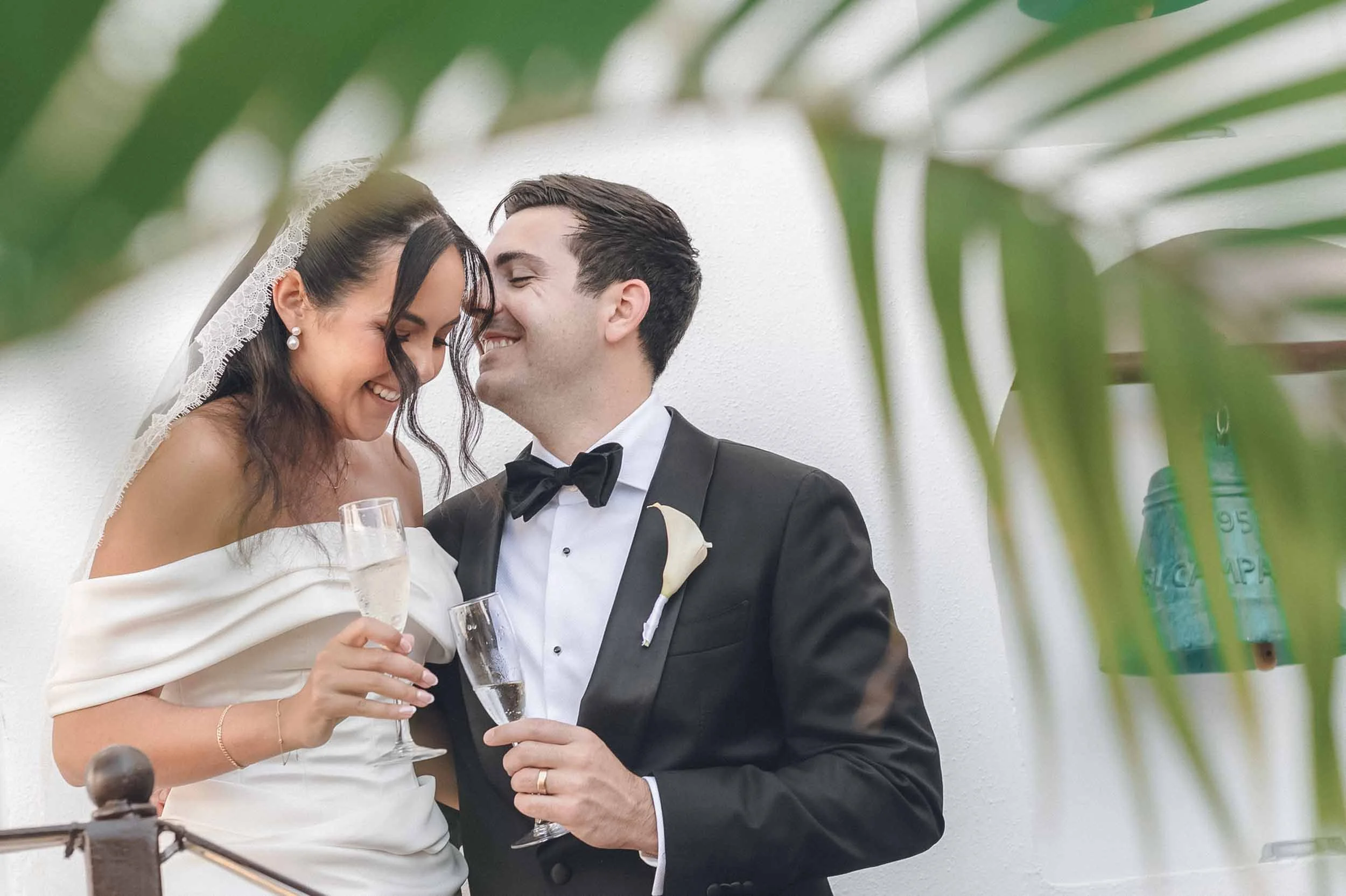 A bride and groom sharing a moment at their wedding, smiling and holding glasses of champagne, partially framed by green leaves.
