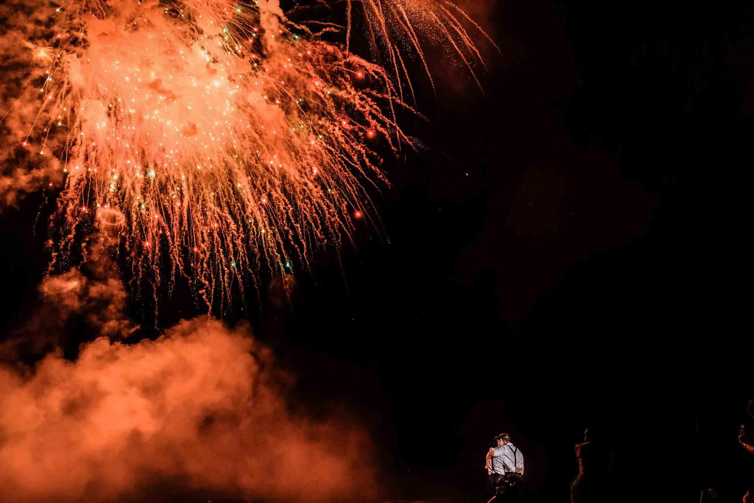 A fireworks display in the night sky with bright orange and red explosions. Two people are standing on the ground watching the fireworks, with one person visible in the lower right corner of the image.
