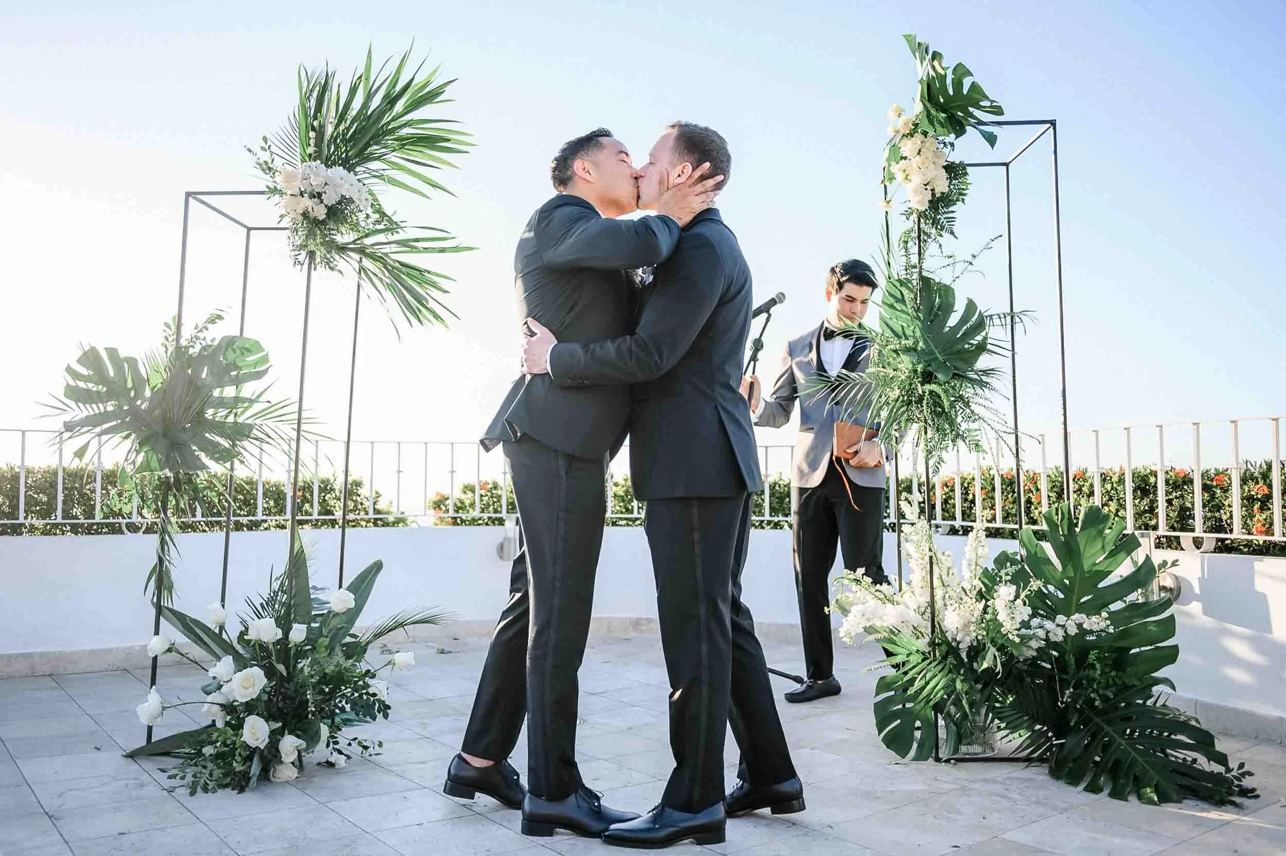 Grooms sharing a kiss at an outdoor wedding ceremony with floral arrangements and green foliage decorations.