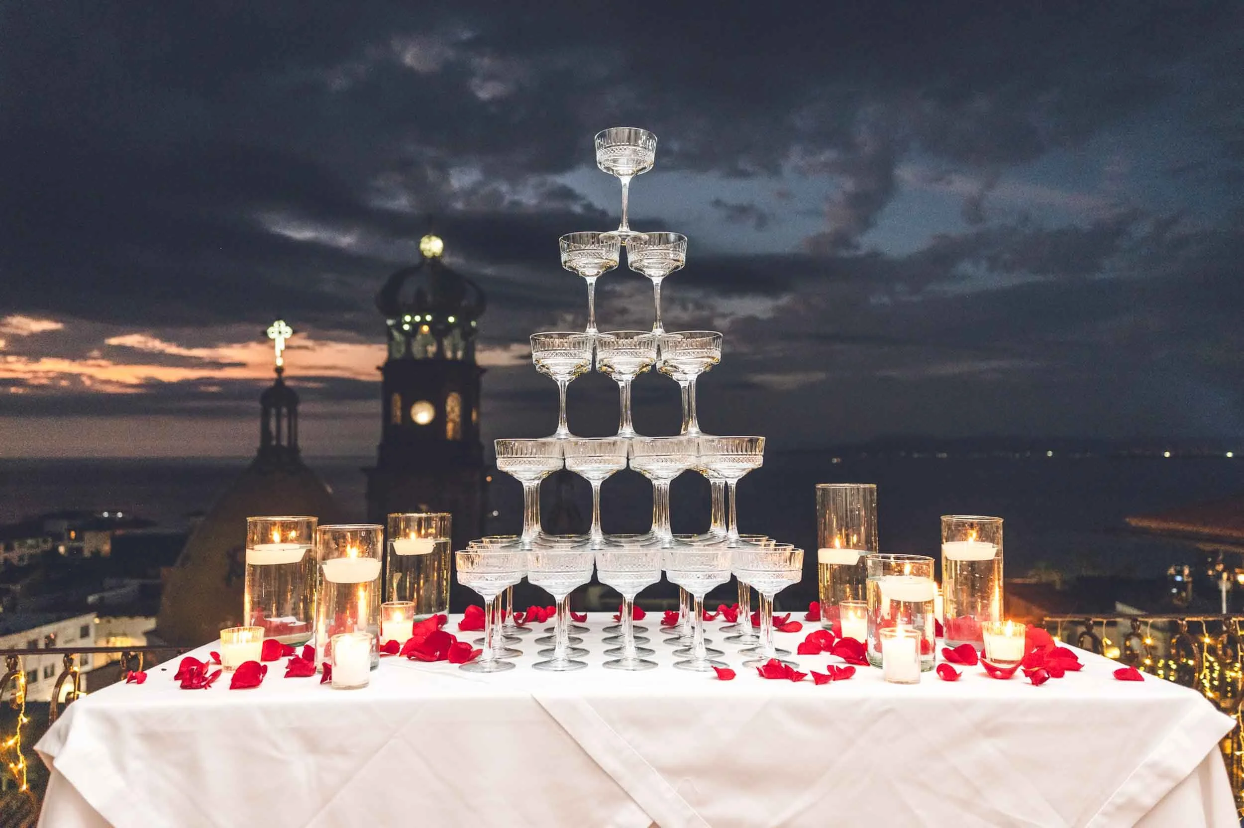 A romantic celebration setup with a pyramid of empty champagne glasses on a table decorated with red rose petals and lit candles, set against a cityscape at dusk with a church tower and dark cloudy sky.