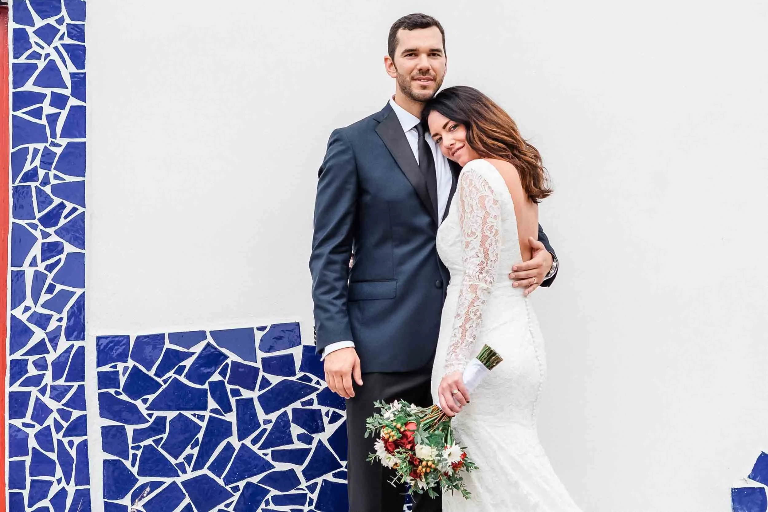 A bride and groom stand close together, smiling at the camera. The groom wears a navy suit with a tie, and the bride is in a white lace wedding dress holding a bouquet of flowers. They are standing in front of a wall with a blue mosaic pattern on the