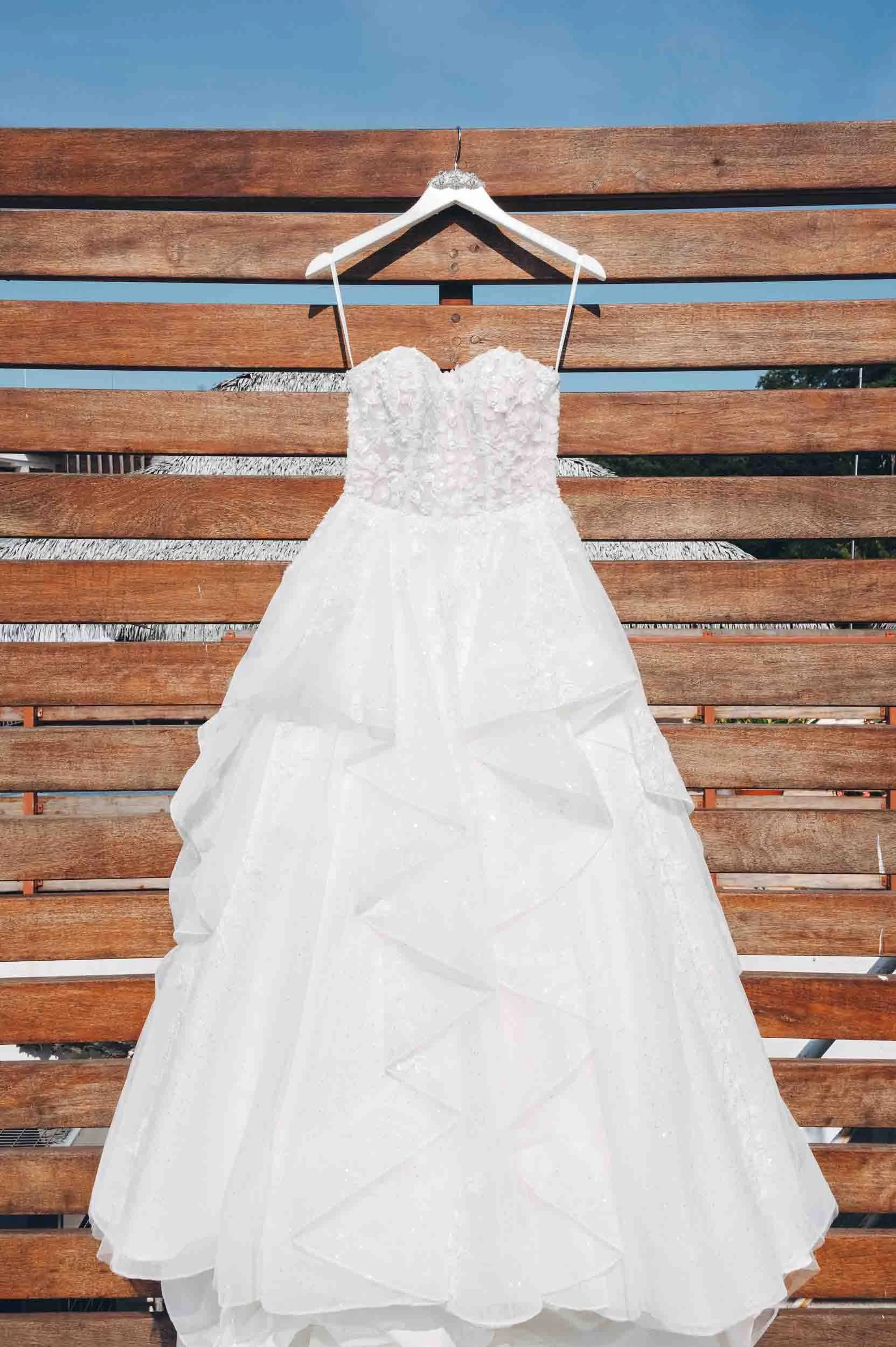 A white wedding dress hanging on a white hanger, displayed against a wooden slat background with blue sky and a tree visible in the distance.