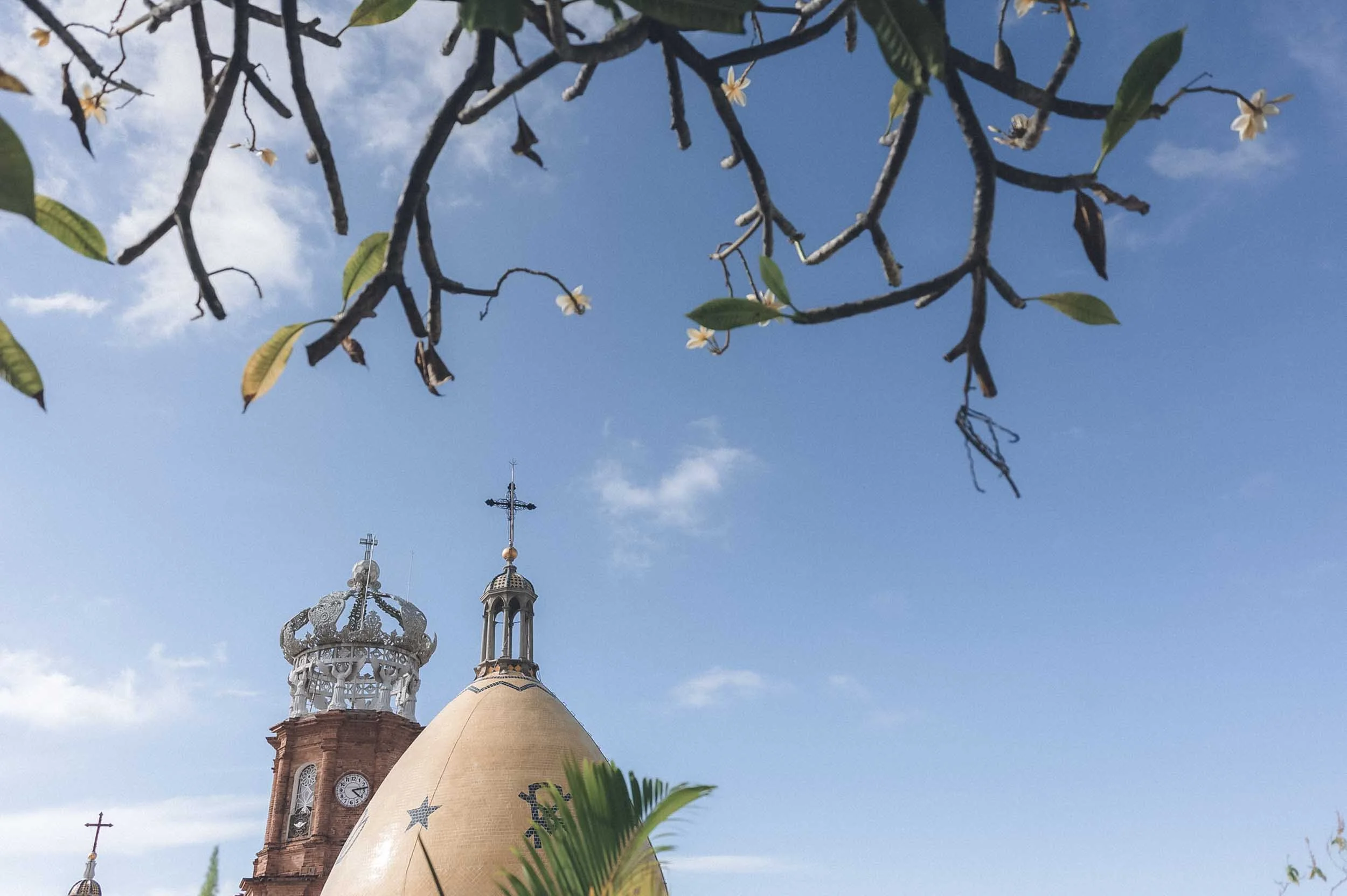 Sky view of a church with two ornate steeples and a domed roof, framed by tree branches and leaves in the foreground, under a partly cloudy sky.