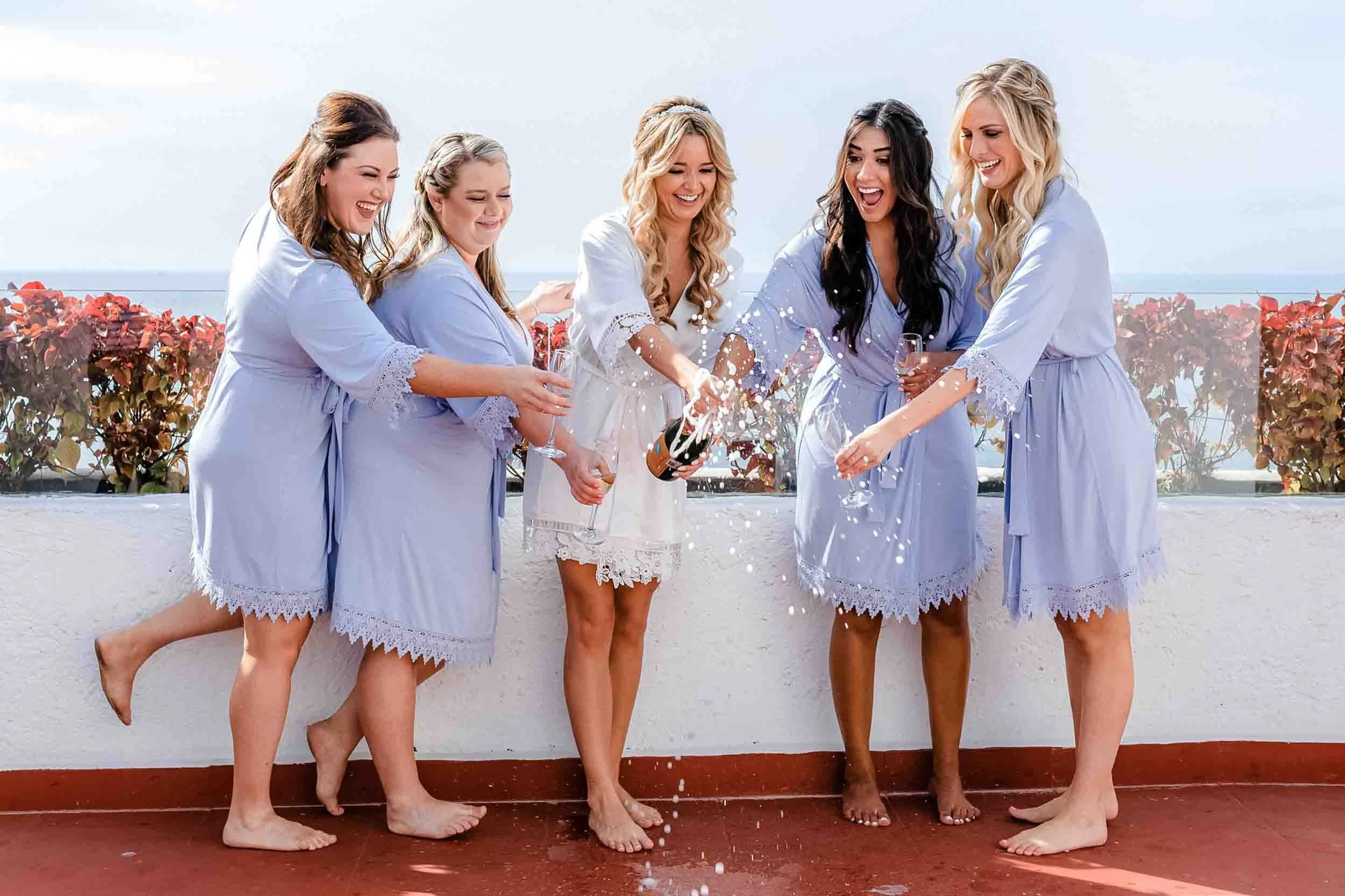 Bride and bridesmaids celebrating with champagne on a balcony overlooking the ocean before the wedding ceremony. 