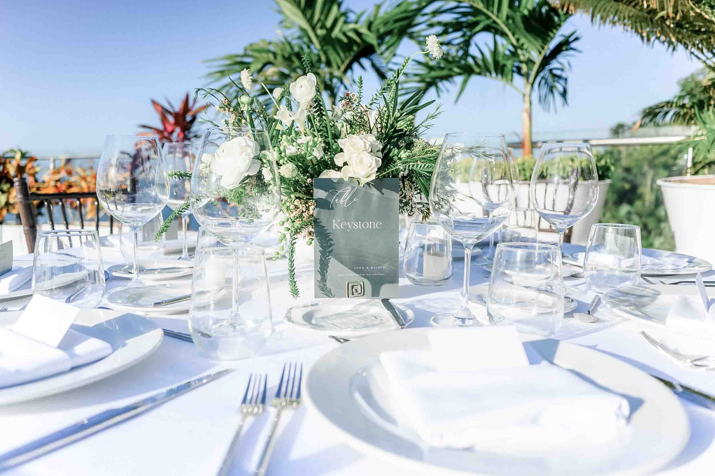 Elegant wedding dining table with a white tablecloth, wine glasses, and a floral centerpiece with white and green flowers on a sunny day, with palm trees in the background.