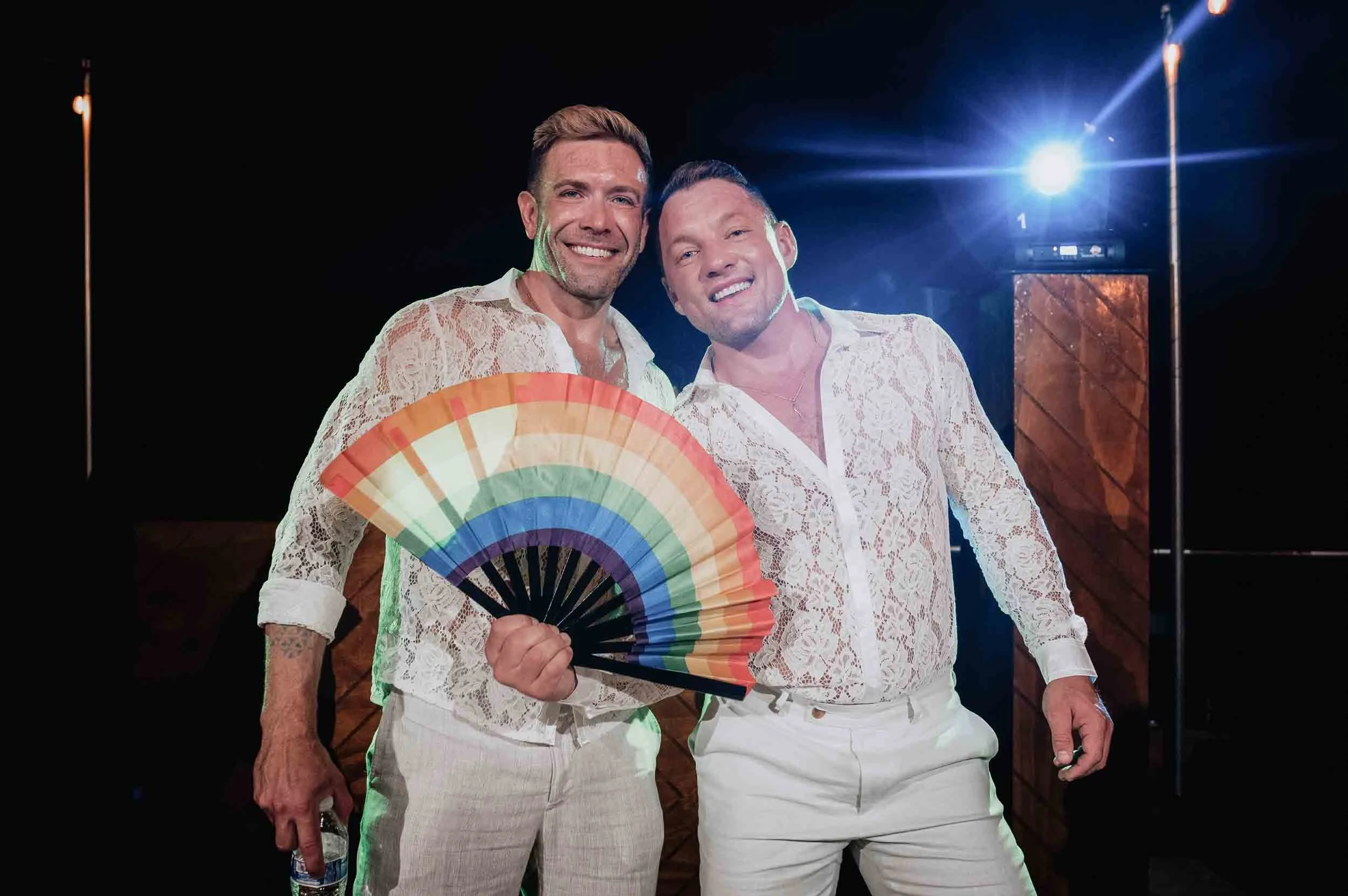 Gay couple in white lace shirts standing close together, one holding a rainbow-colored fan, having fun at their wedding reception.