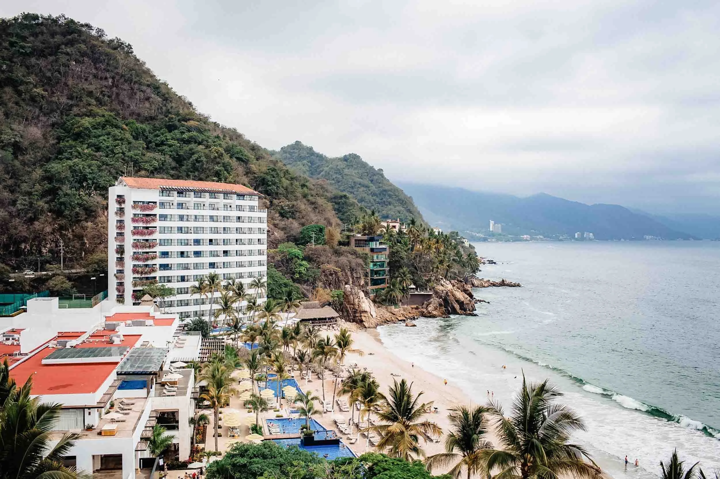 View of the Hyatt Ziva seaside resort with a sandy beach, palm trees, and a tall white hotel building with red shutters. There are swimming pools and umbrellas on the beach, and mountains covered with greenery in the background under overcast sky.